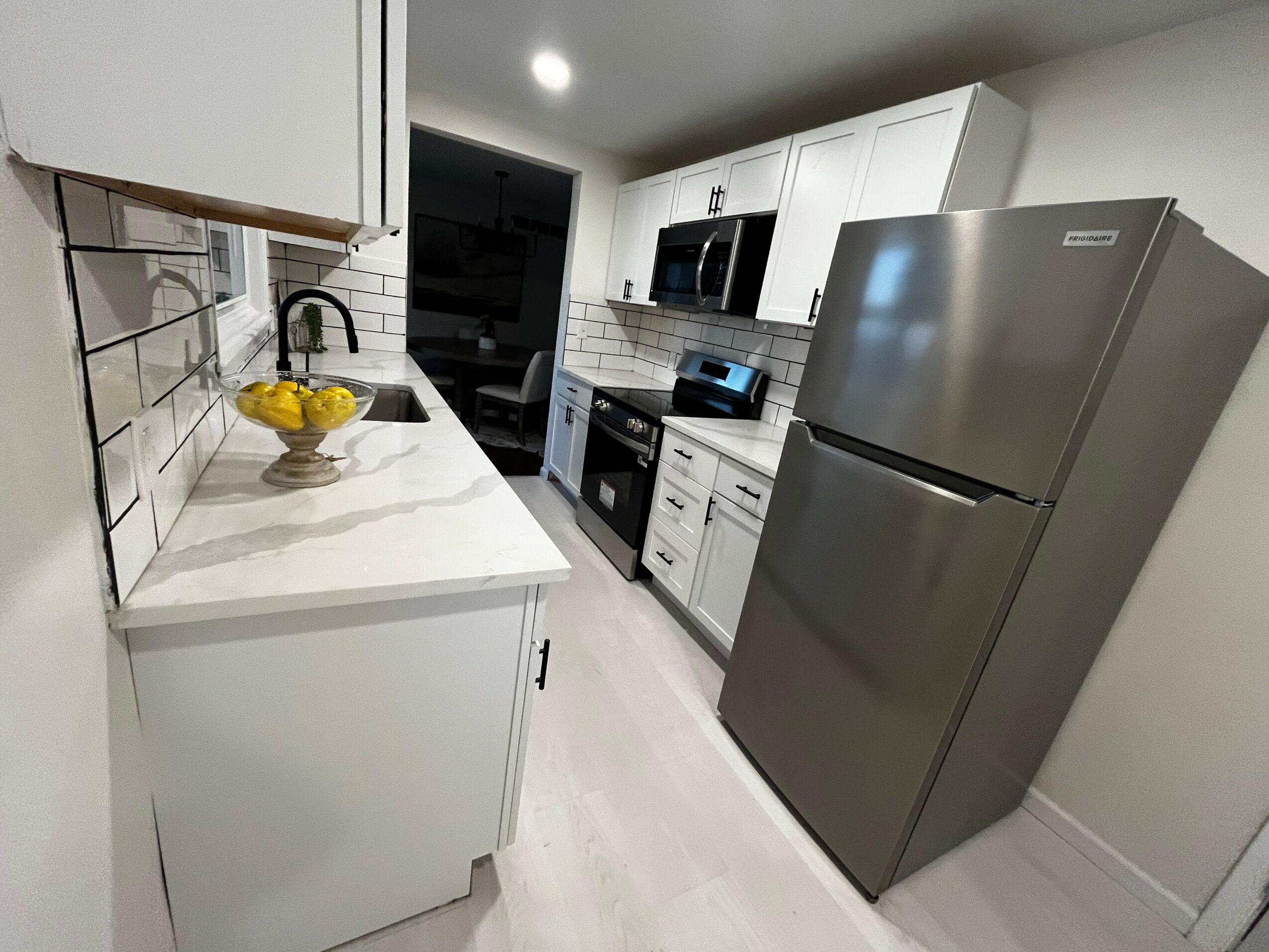 Modern kitchen with white cabinets, black appliances, and a stainless steel refrigerator, viewed from the doorway. Bright lighting and tiled backsplash.