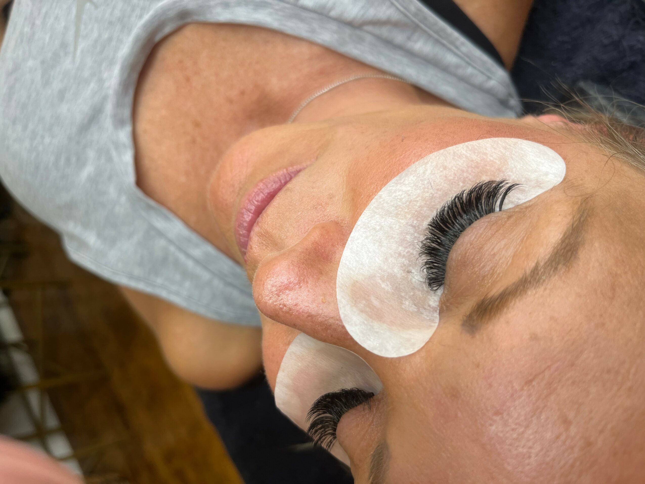 Close-up of a woman with eye patches and eyelash extensions, lying down with her eyes closed.