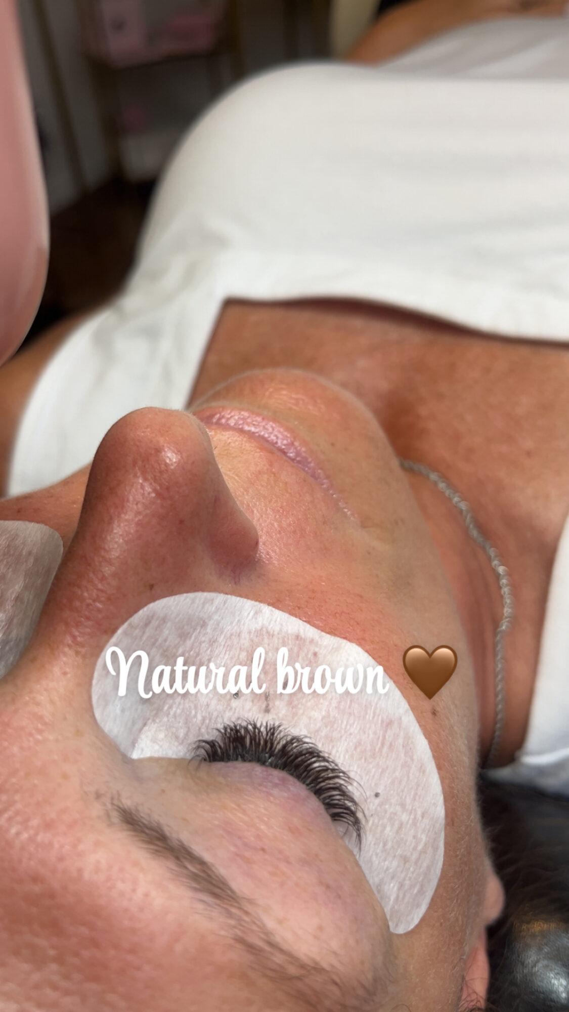 Close-up of a person's face with a white eye patch labeled 'Natural brown' and a brown heart icon, lying on a bed.