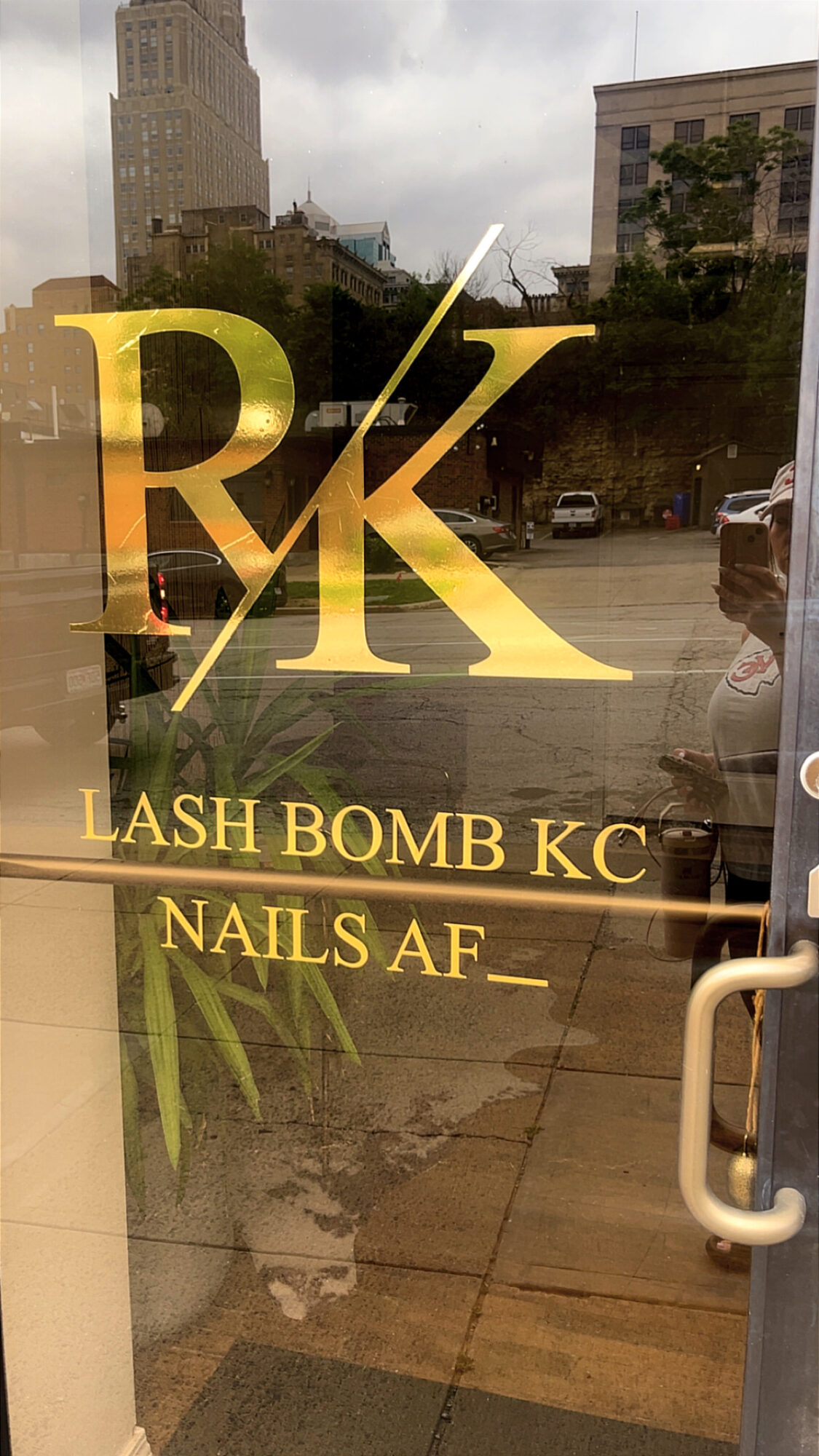 Store window with large gold letters 'RK' and text 'LASH BOMB KC NAILS AF' below, reflecting street scene.