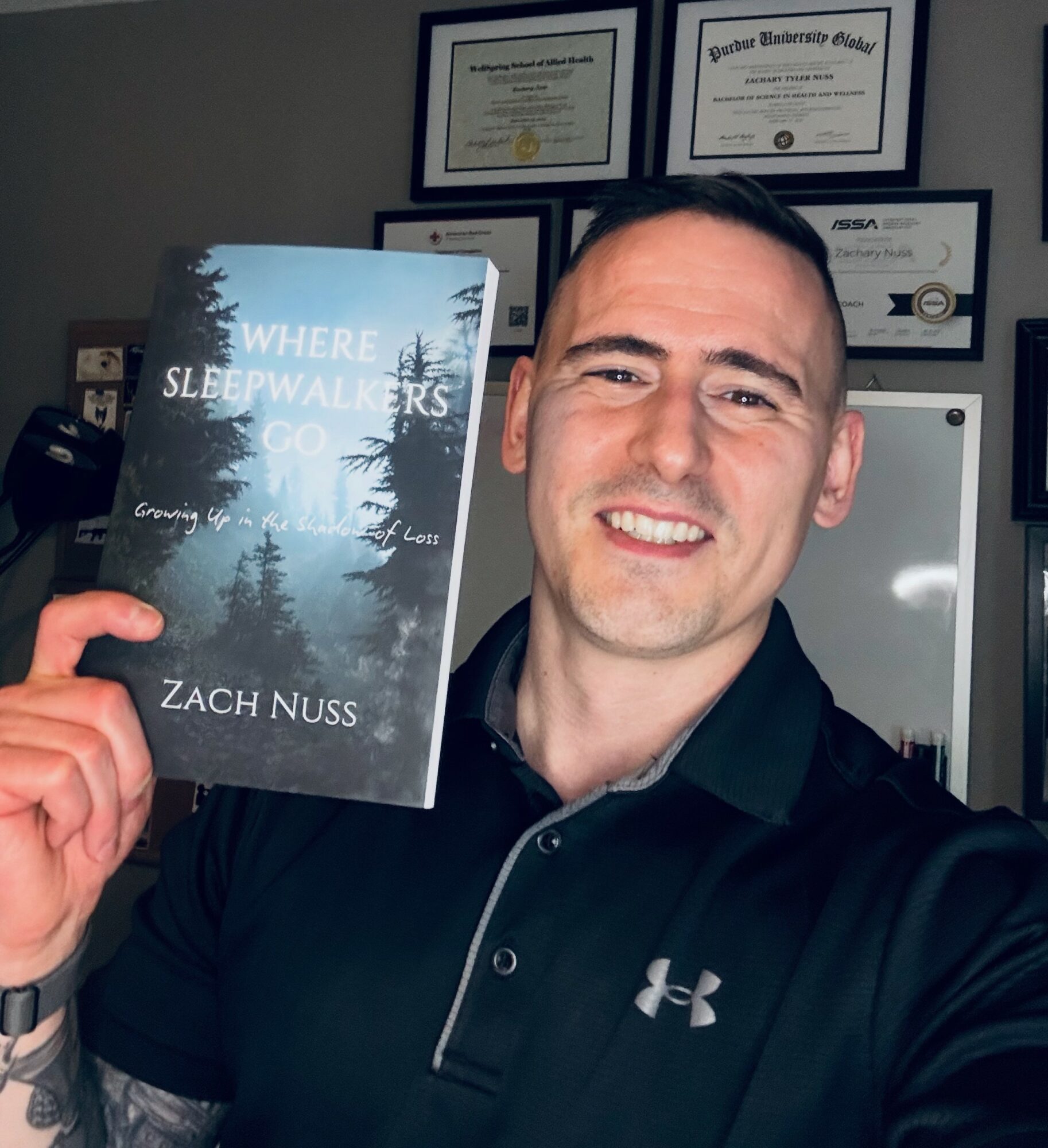 Man holding a book titled 'Where Sleepwalkers Go' by Zach Nuss, smiling, certificates on wall behind him.