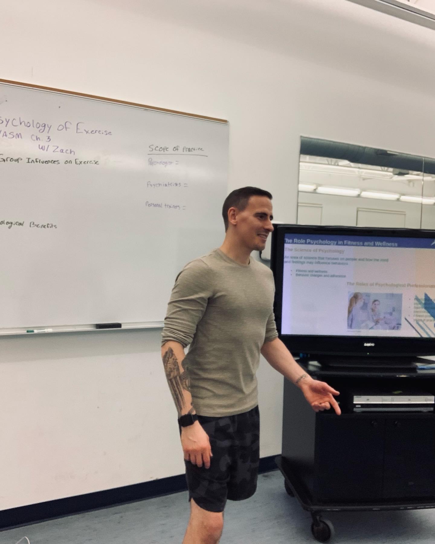 Man standing in front of a whiteboard and a large screen, gesturing with his right hand, in a classroom setting.