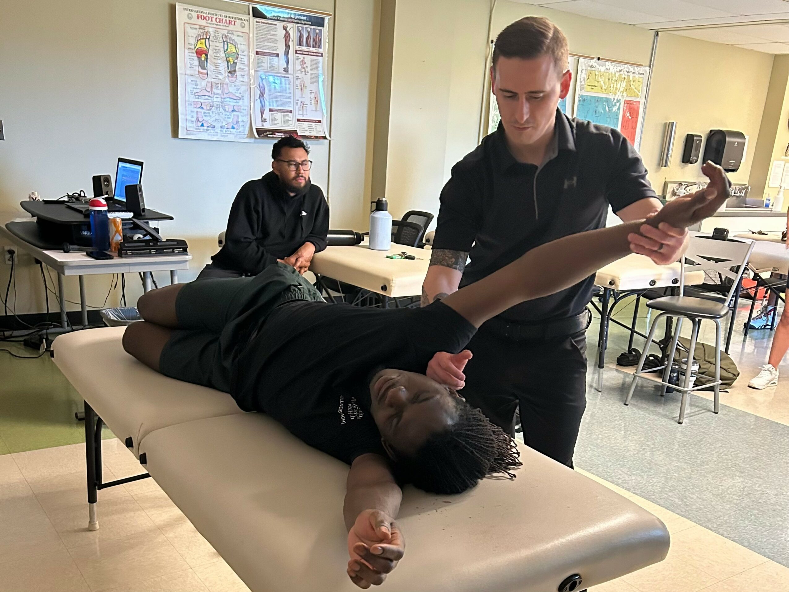 A person lying on a medical examination table with an arm extended, being attended to by a healthcare worker in a clinical setting.