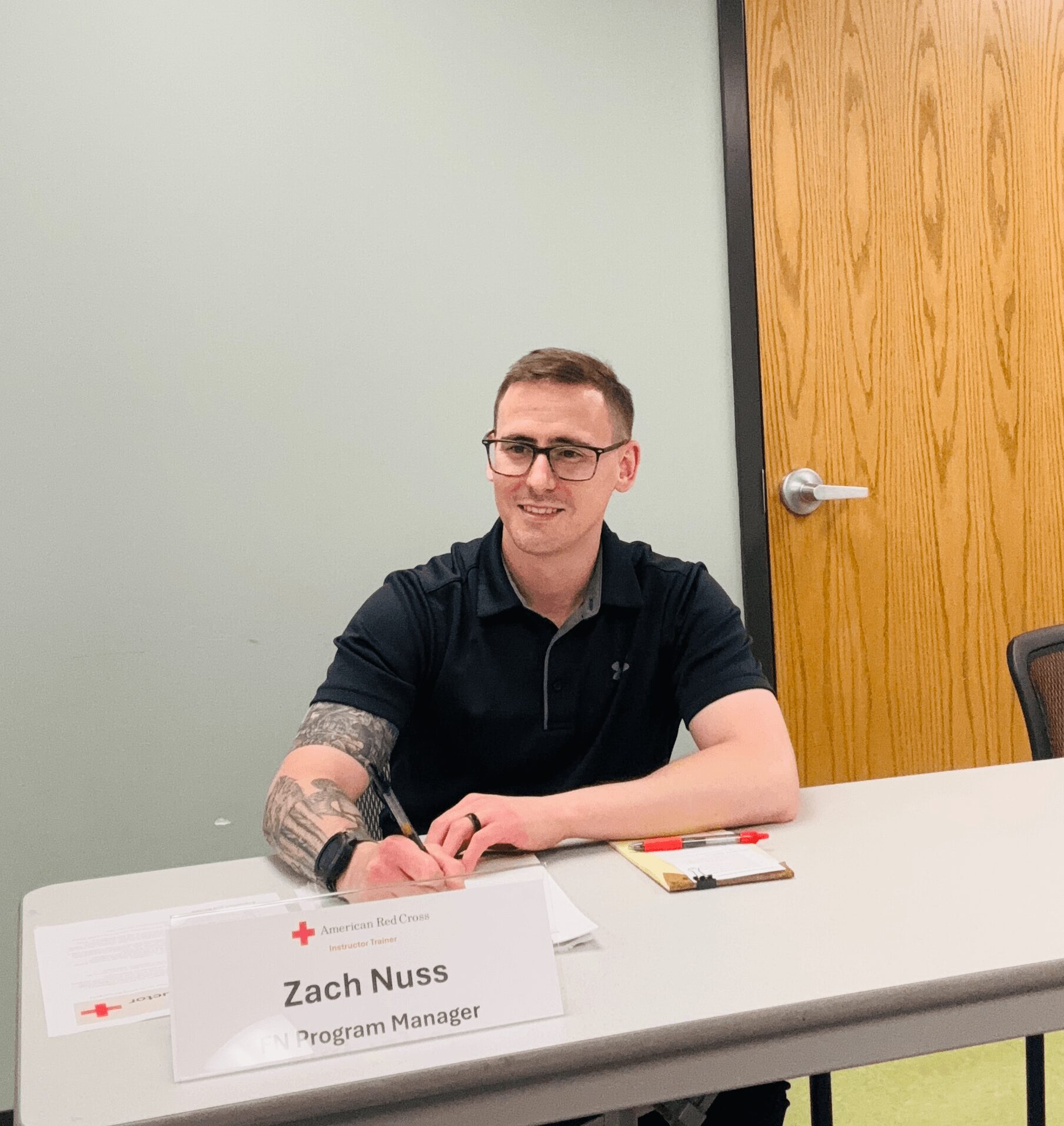 Young man with glasses and tattoos sitting at a table, smiling, with a nameplate reading Zach Nuss, Program Manager.