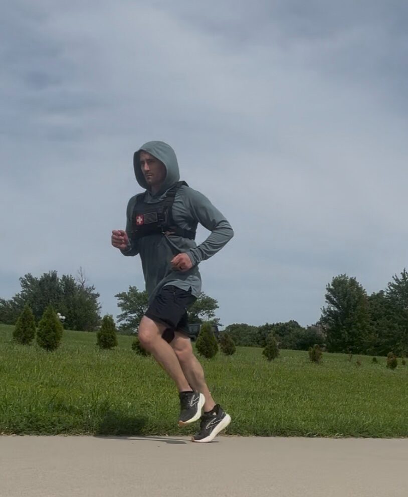 Person running outdoors on a cloudy day, wearing a hoodie, jacket, shorts, and running shoes, with trees and grass in the background.