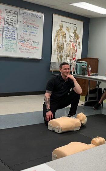 Man kneeling on floor next to CPR mannequins in training room with medical chart and skeleton diagram on wall.