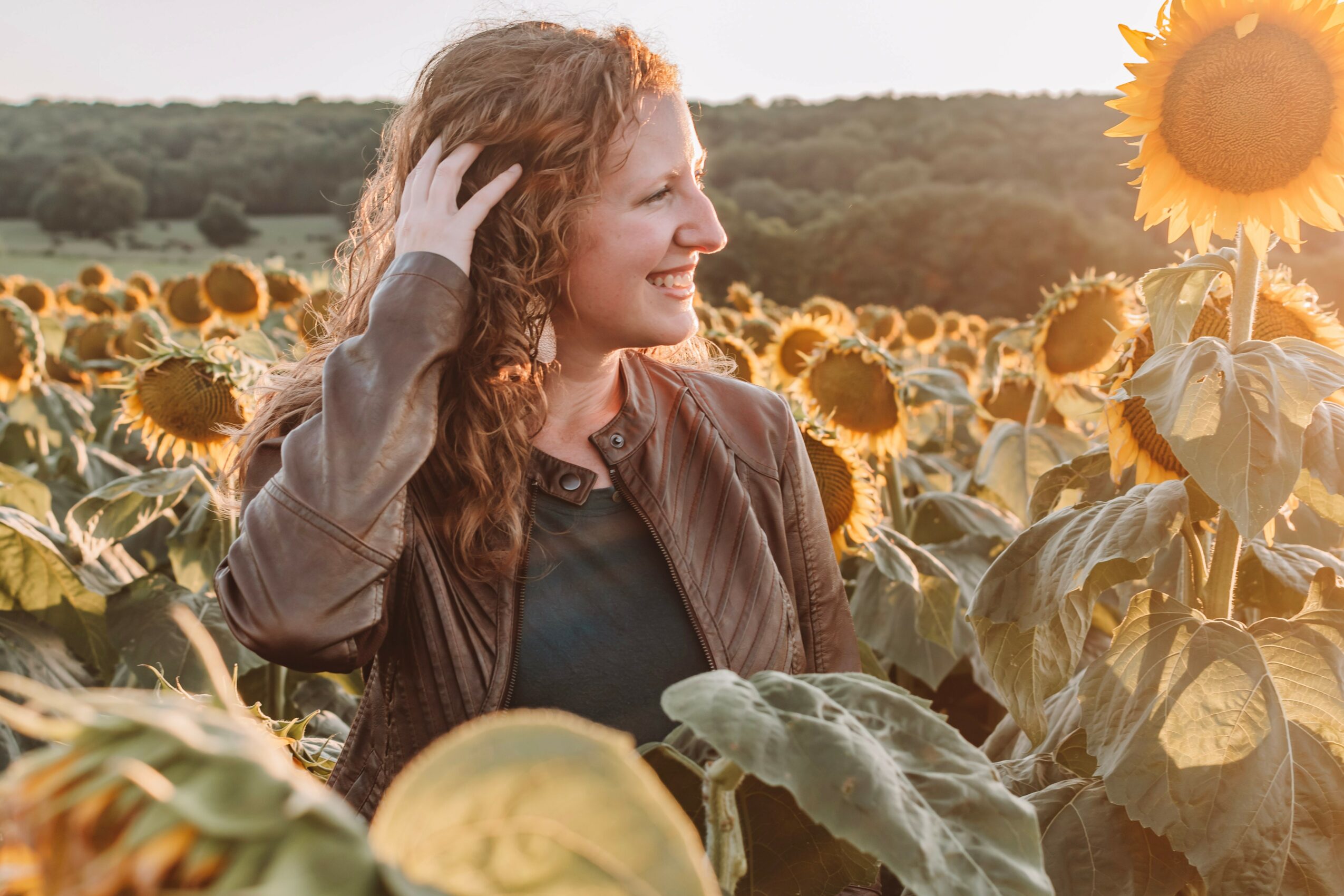 Woman with curly hair smiling in a sunflower field during sunset, wearing a brown jacket and black shirt.