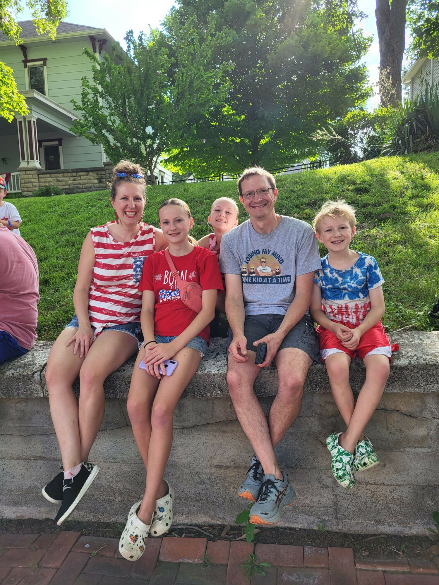 Four people sitting on a brick ledge outdoors, surrounded by green trees and grass, smiling at the camera.