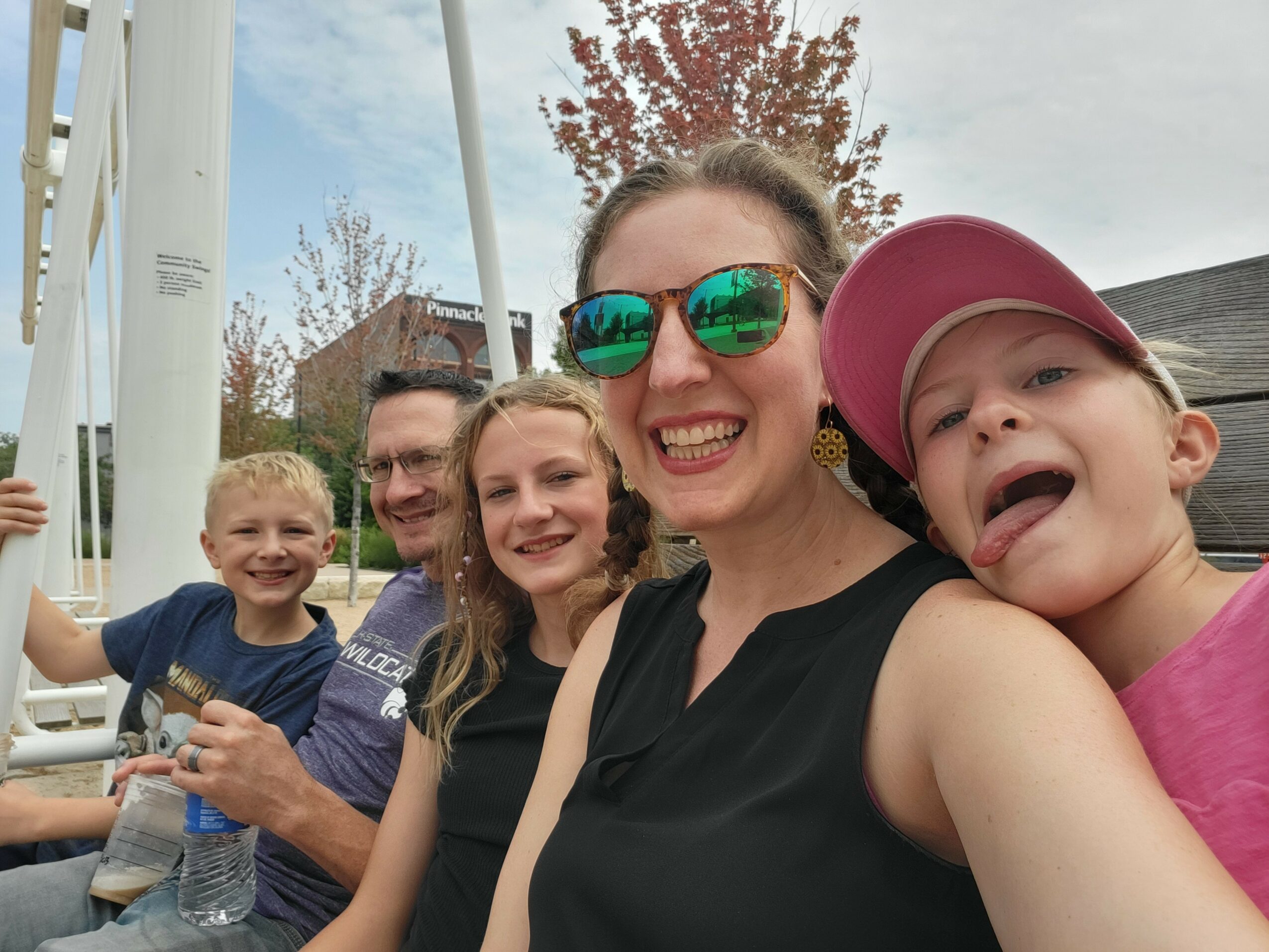 Group of five smiling people, including children and adults, outdoors on a sunny day, sitting together and taking a selfie.
