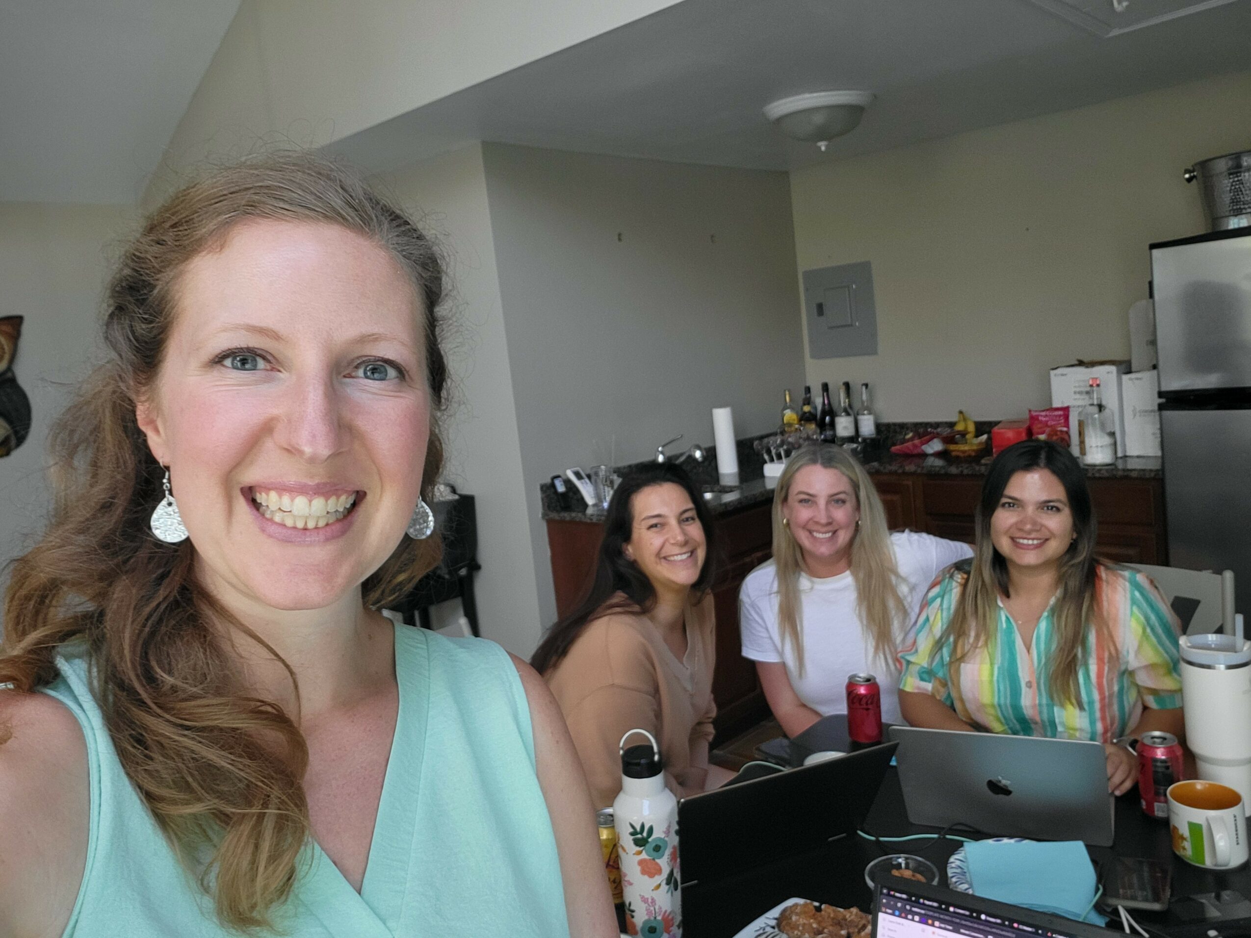 Four women smiling at a table with a laptop and drinks in a room with kitchen appliances.