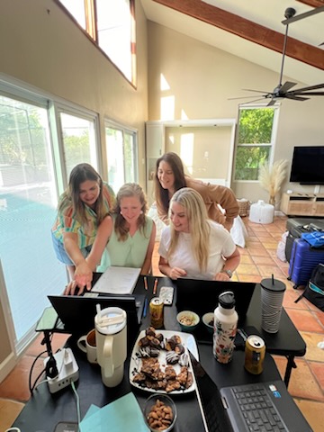 Four women gathered around a table with food and drinks in a bright room with large windows and a ceiling fan.