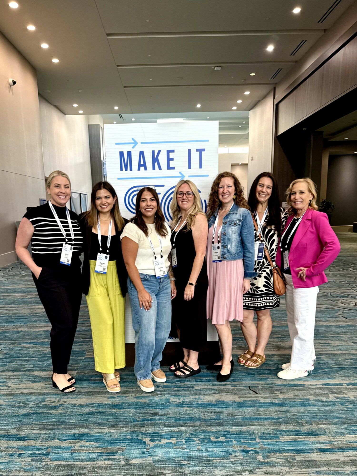 Group of six women standing in front of a screen with 'MAKE IT' text, smiling indoors at a conference or event.