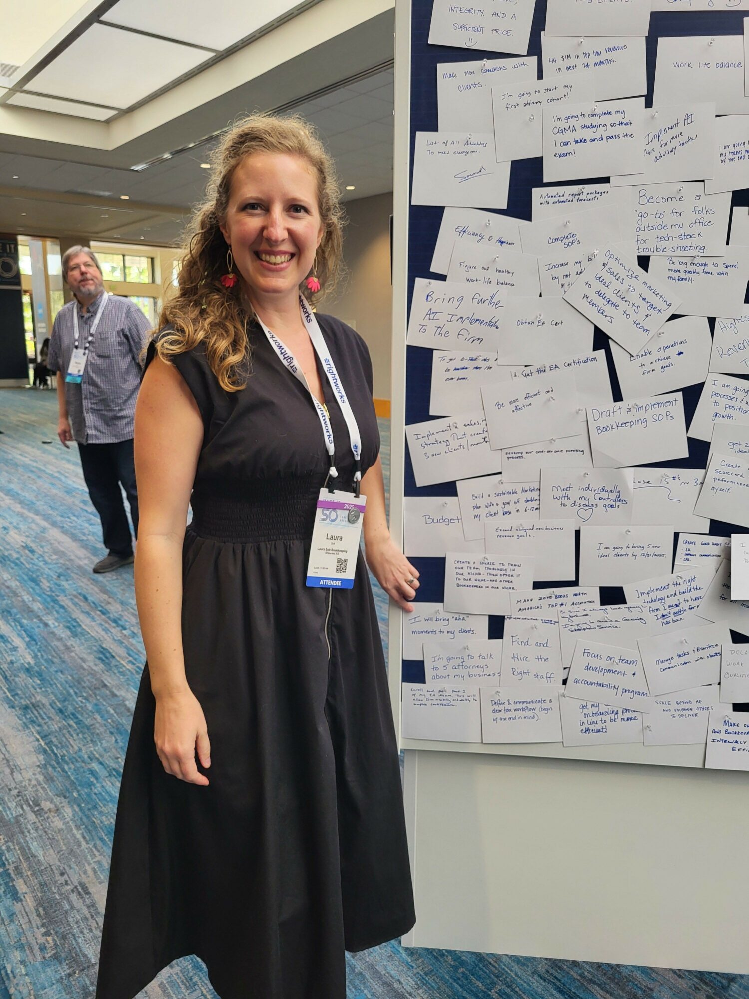 Woman in black dress standing in a room with notes on a board behind her, smiling, with a lanyard and badge.