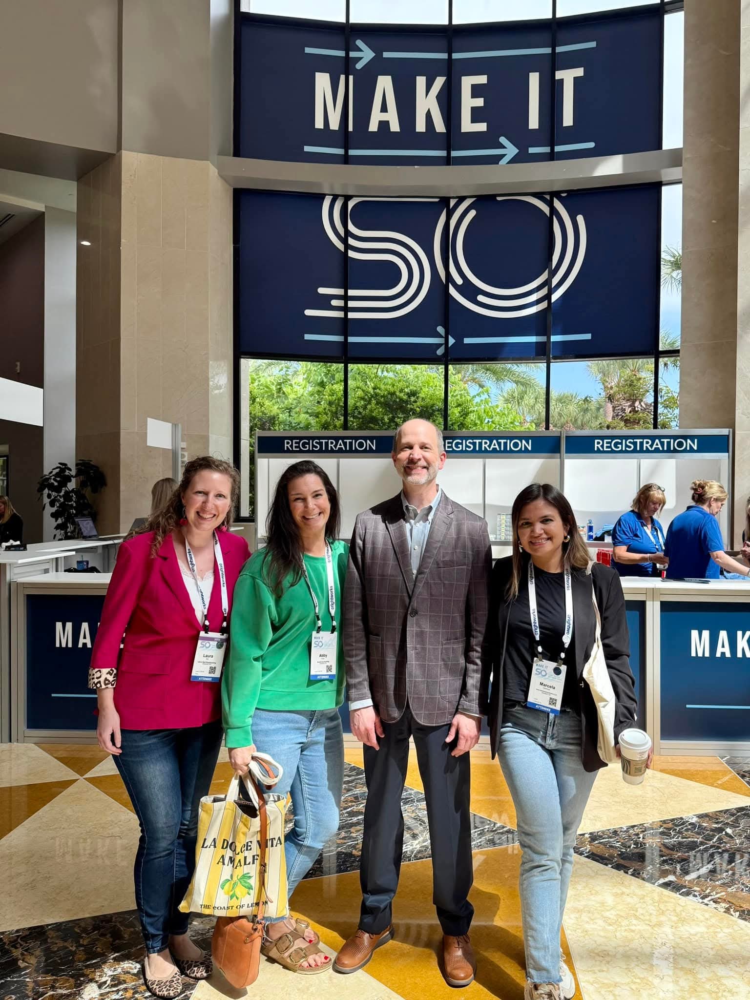 Four people standing in front of registration desks at a conference, smiling, with a large sign that says 'MAKE IT SO' behind them.
