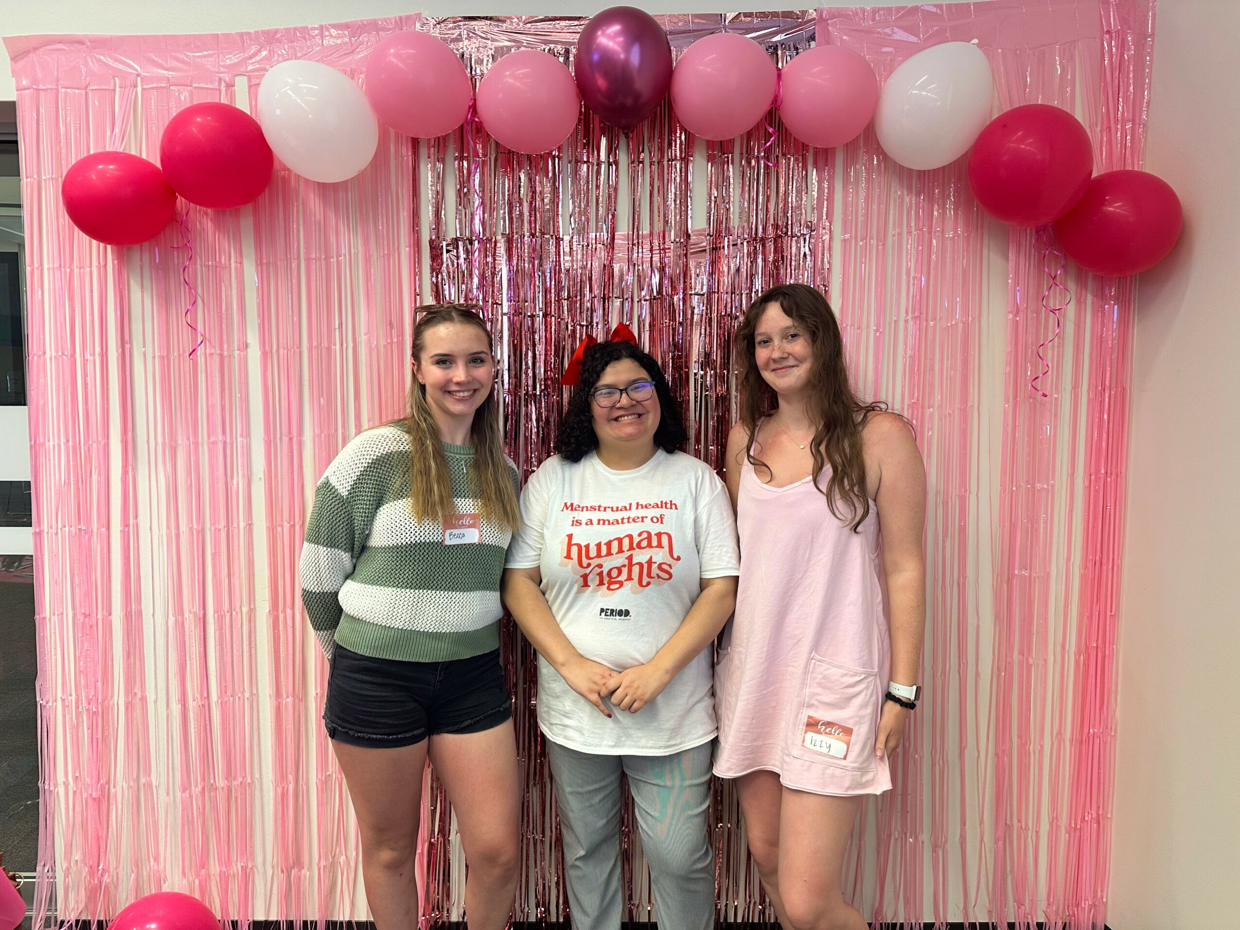 Three women stand in front of pink and purple balloons and streamers, smiling at the camera.