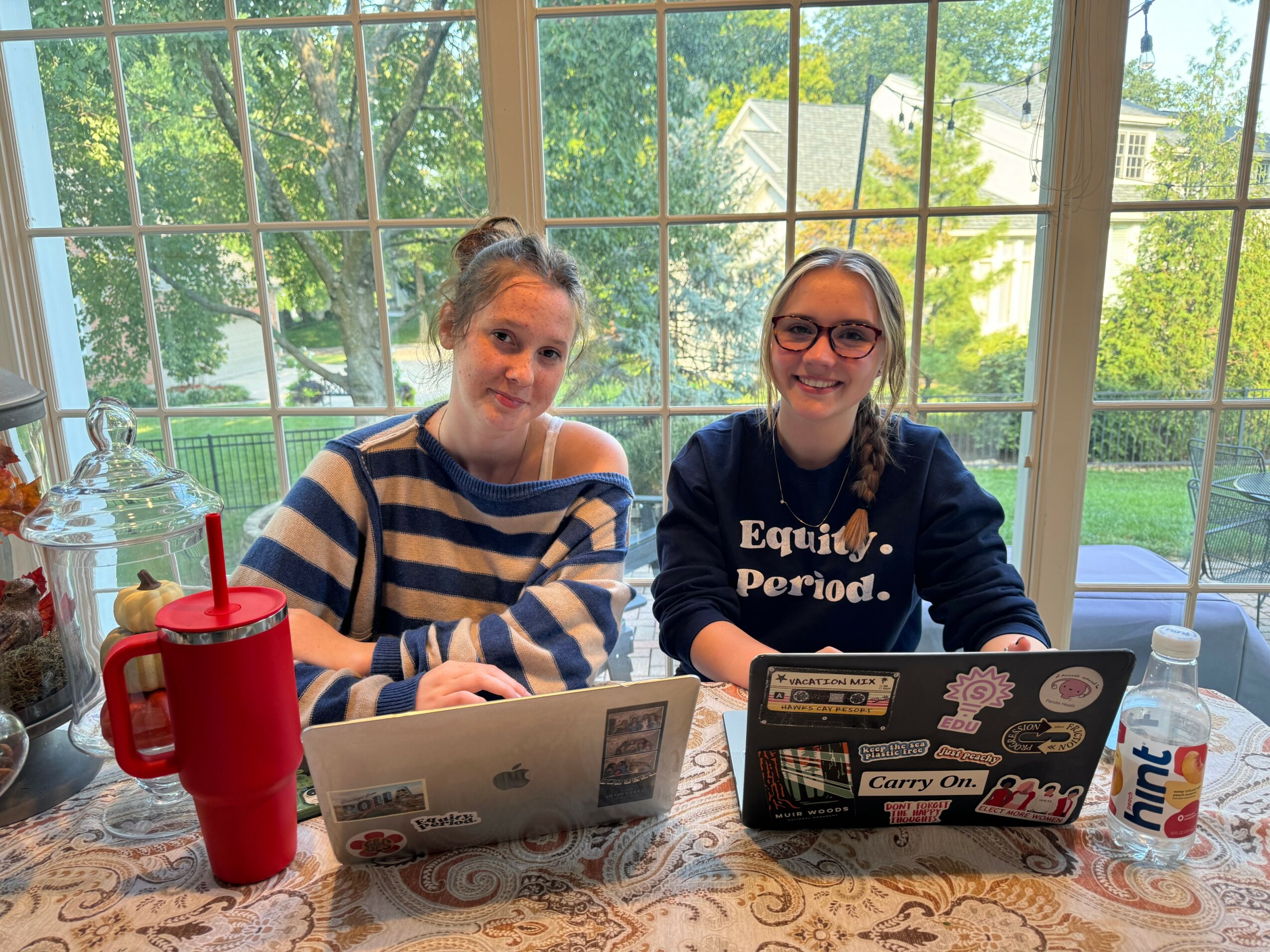 Two girls sit at a table with laptops, a red water bottle, and a can of Fanta, in front of large window.