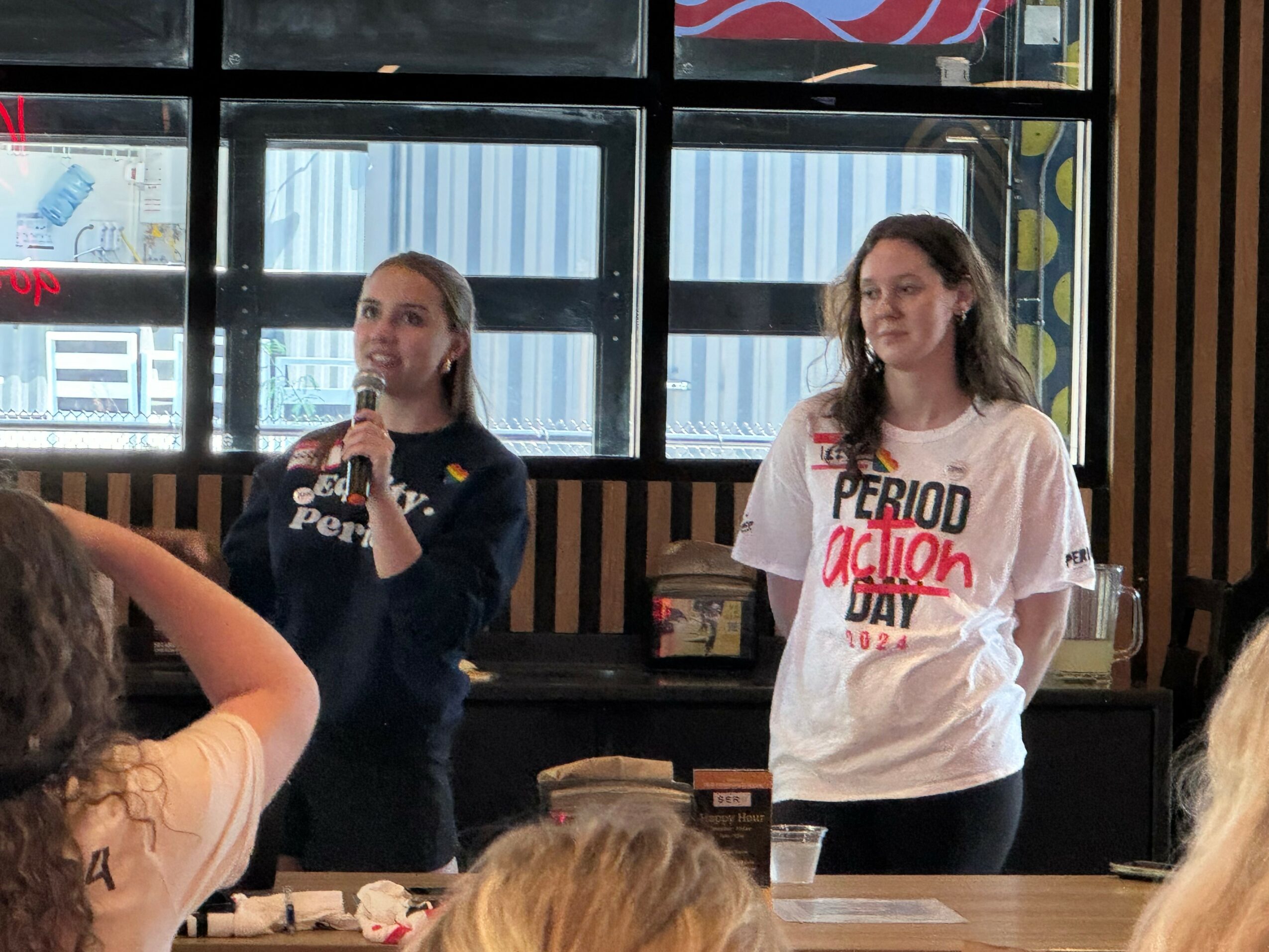 Two women stand in front of a large window, speaking to a group of people in a room with wooden walls.