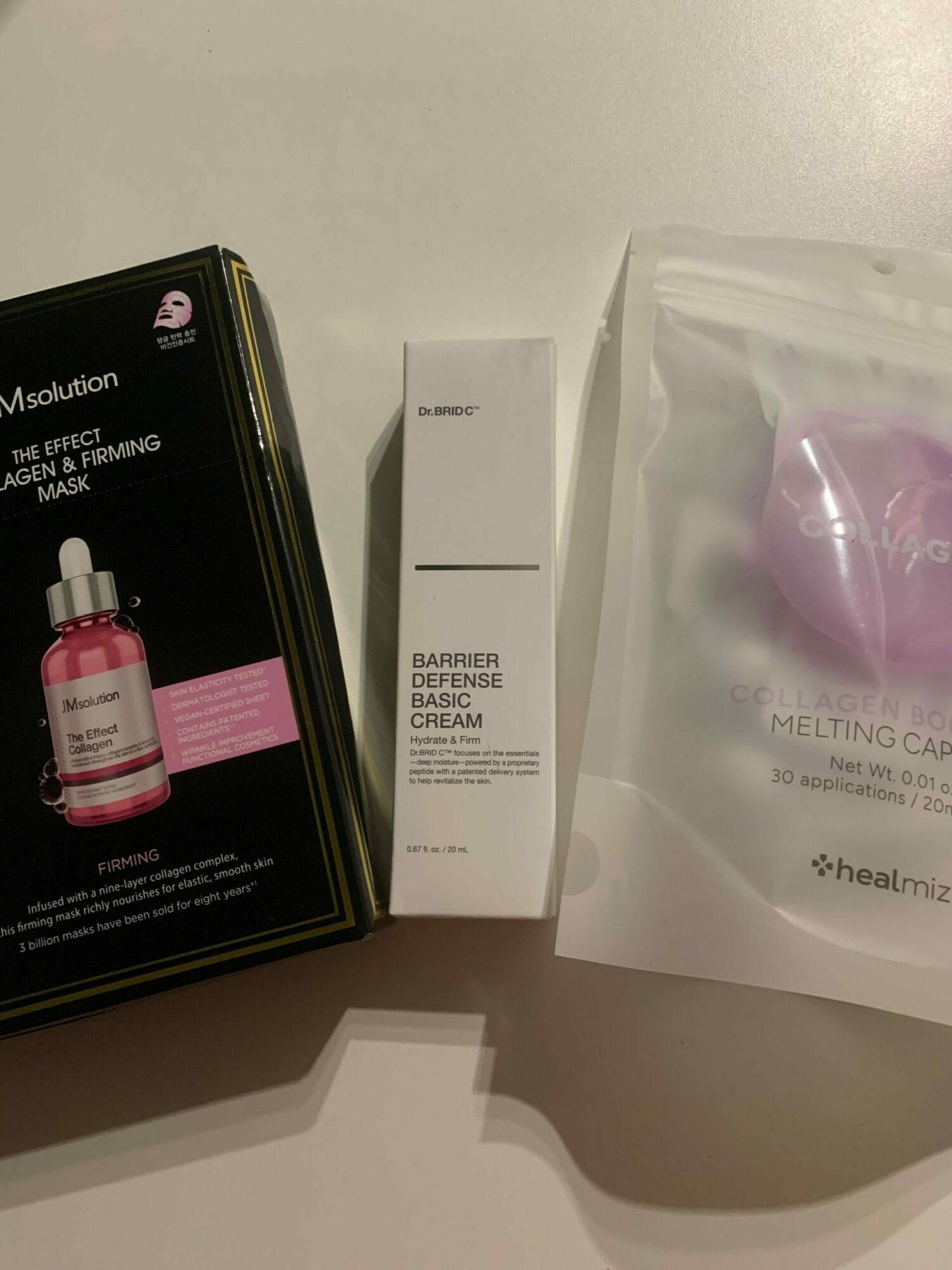 Various skincare products, including a pink dropper bottle, a white box, and a clear bag with purple items, on a white surface.