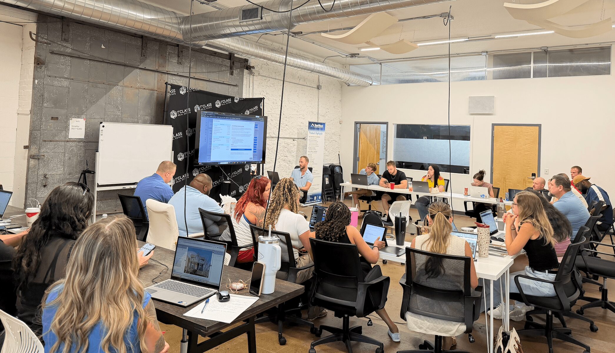 People seated at tables in a classroom or conference room with screens and laptops, listening to a presentation.