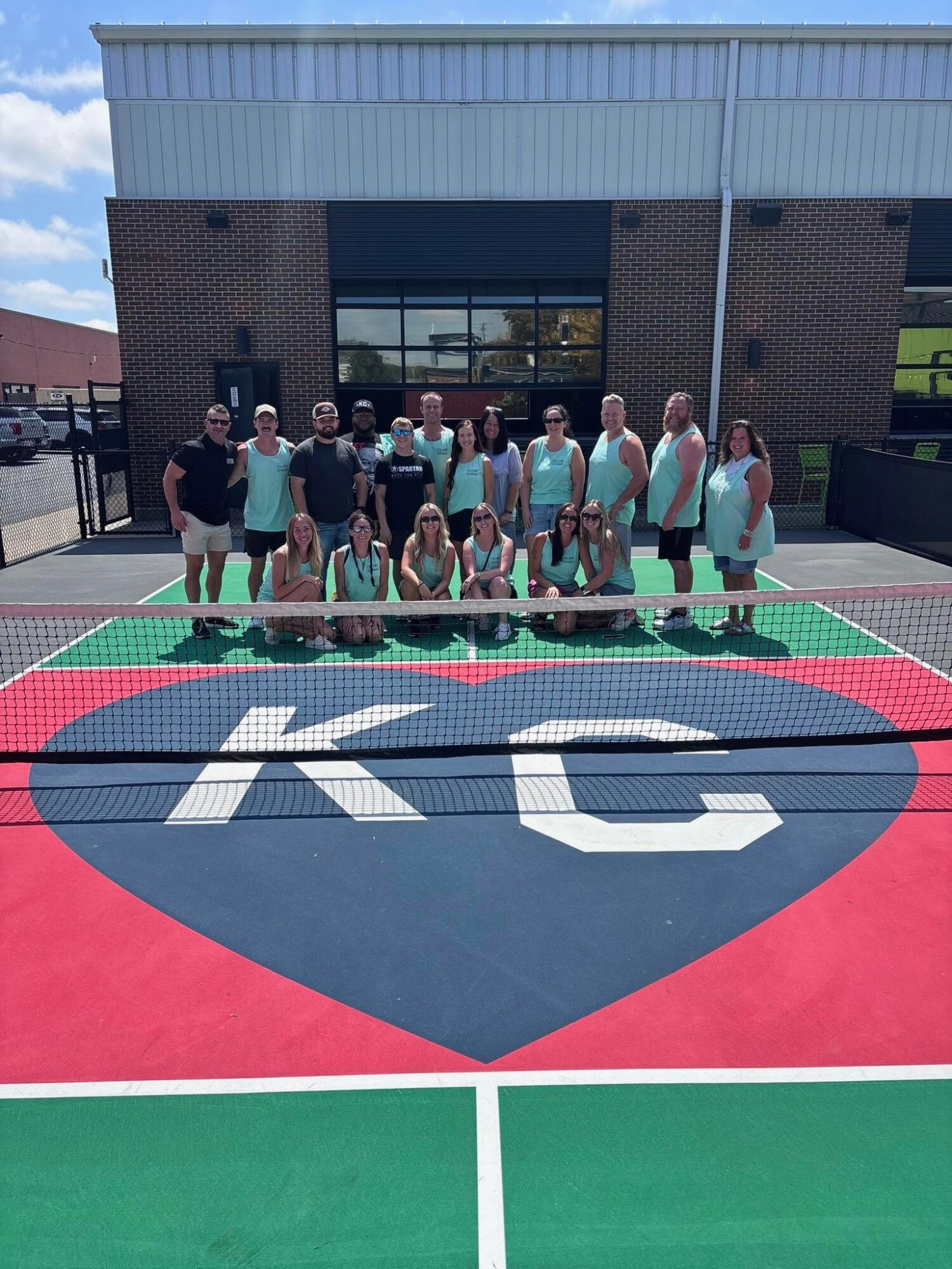 Group of people standing and kneeling on a colorful court with a large logo, in front of a building with glass windows.