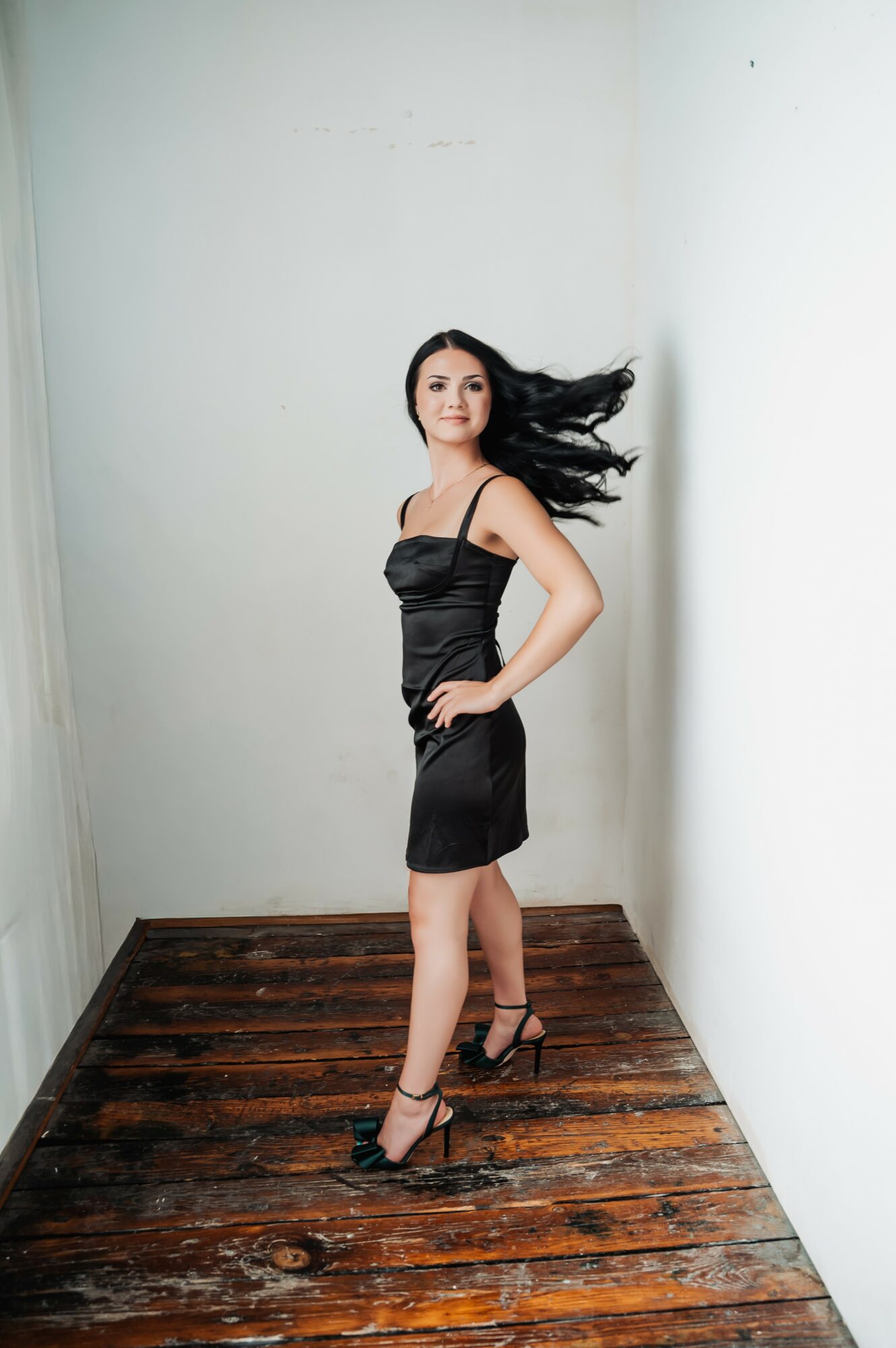 Woman in black dress standing on wooden floor with hair flowing, white walls in background.