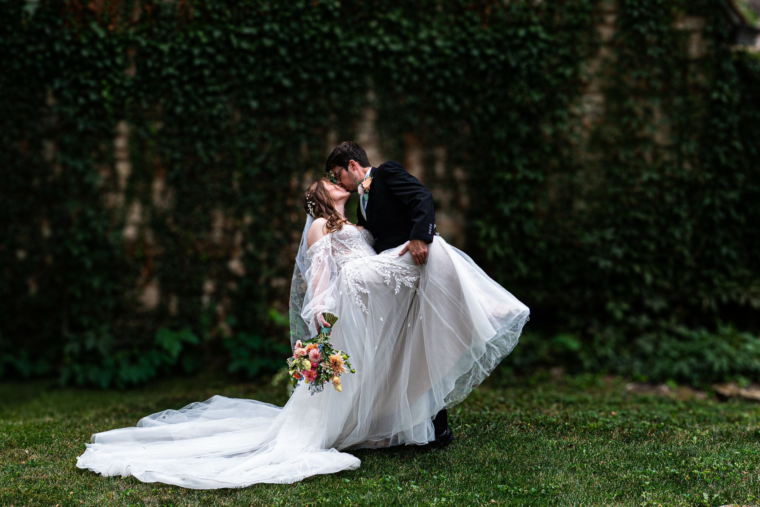 A bride and groom share a kiss outdoors, with the groom lifting the bride's dress, surrounded by greenery.
