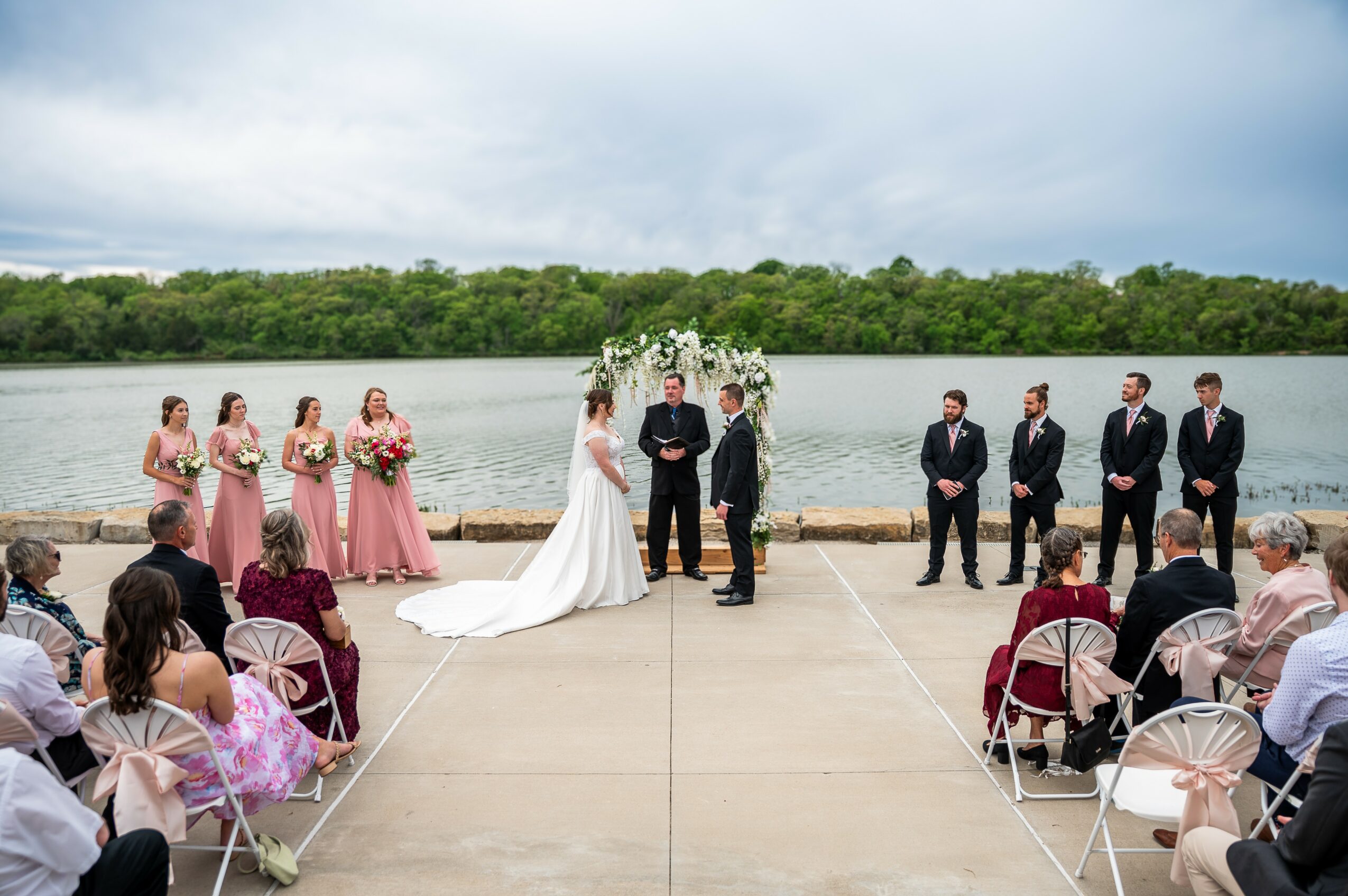 Wedding ceremony outdoors by water with bride, groom, officiant, bridesmaids, groomsmen, and seated guests, overcast sky.
