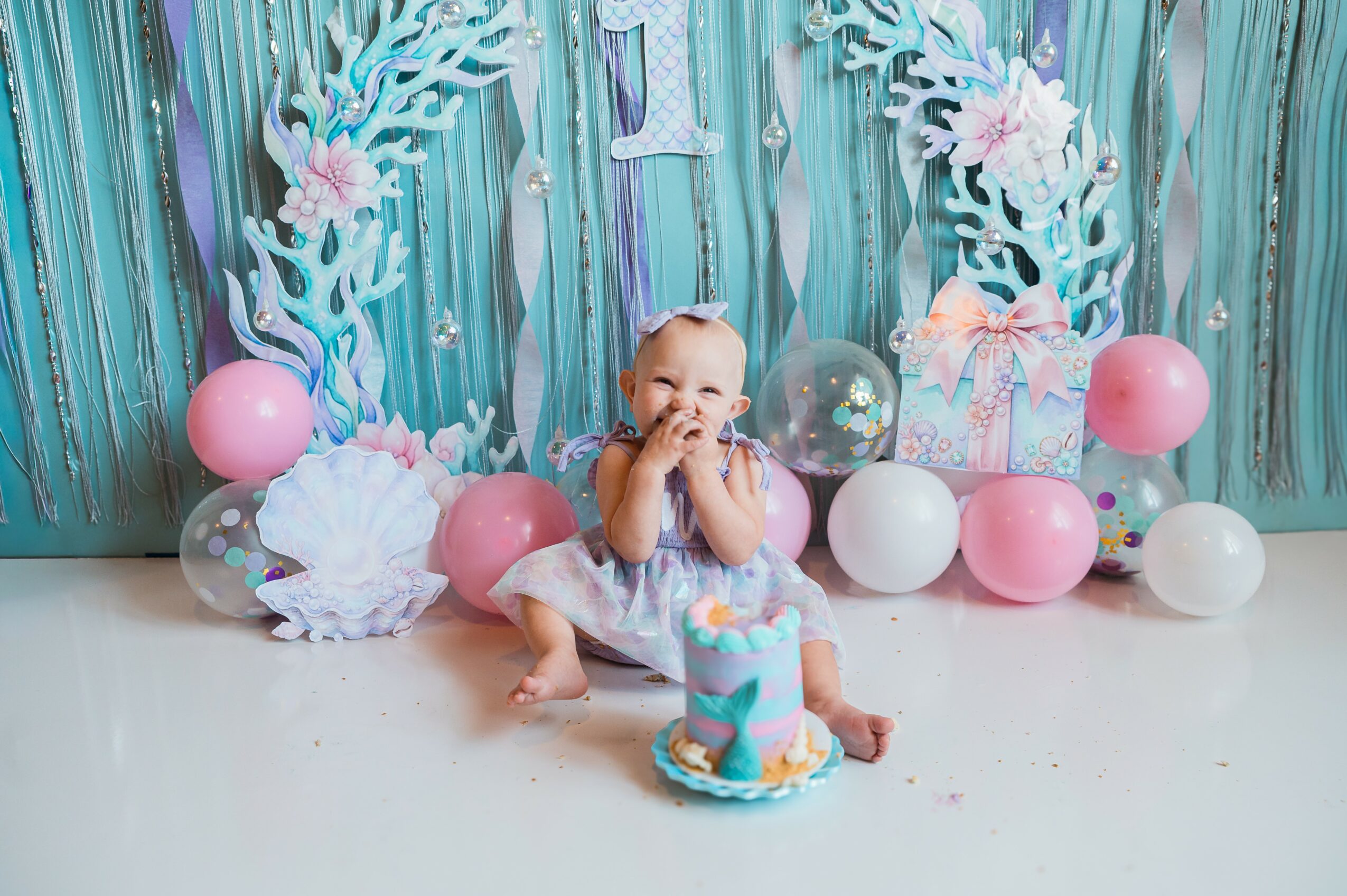 Baby sitting in front of a birthday cake with decorations including pink and white balloons, a gift box, and a backdrop with blue and white decorations.