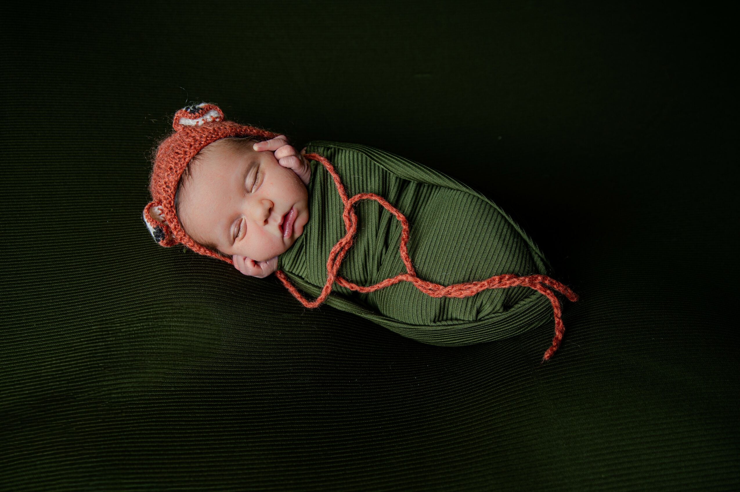 Newborn baby sleeping wrapped in green fabric, wearing a red knit hat with a pom-pom, on a dark background.