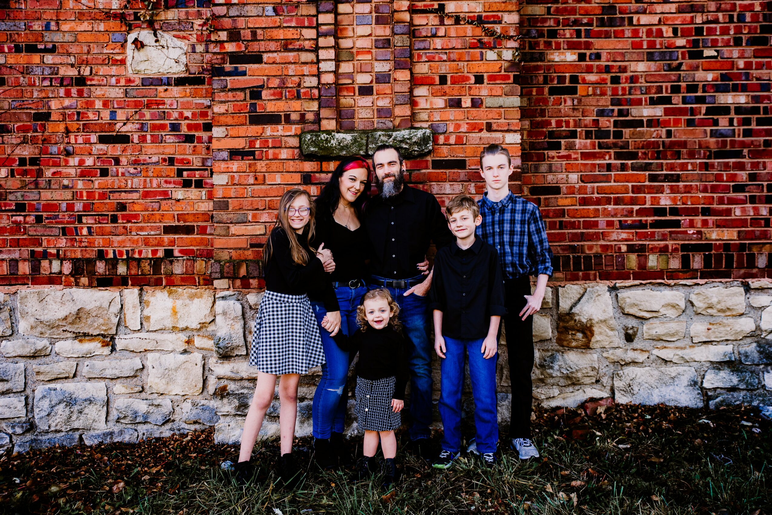 Group of six people standing outdoors in front of a brick wall, smiling, with grass and soil at their feet.