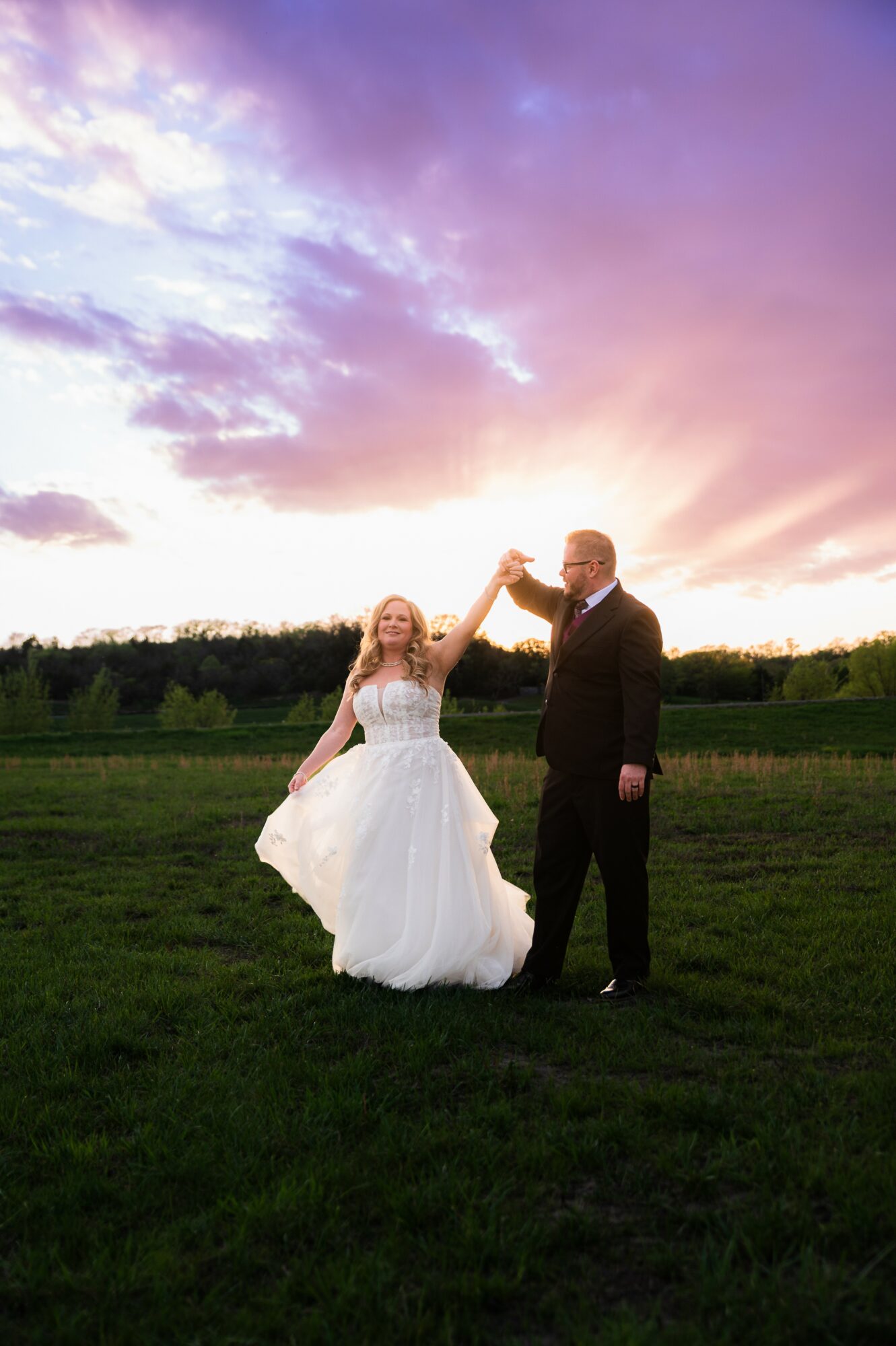 A bride and groom dance outdoors at sunset, holding hands, with a colorful sky and green field in the background.