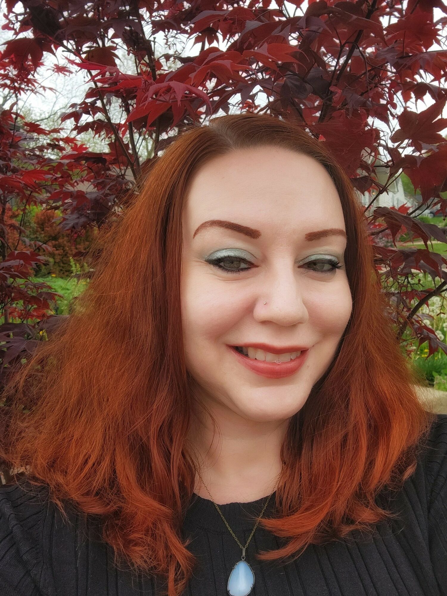 Woman with red hair smiling outdoors with red and green foliage in background.