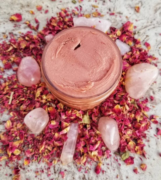 Pink cream in a jar surrounded by rose petals and small stones on a textured surface.