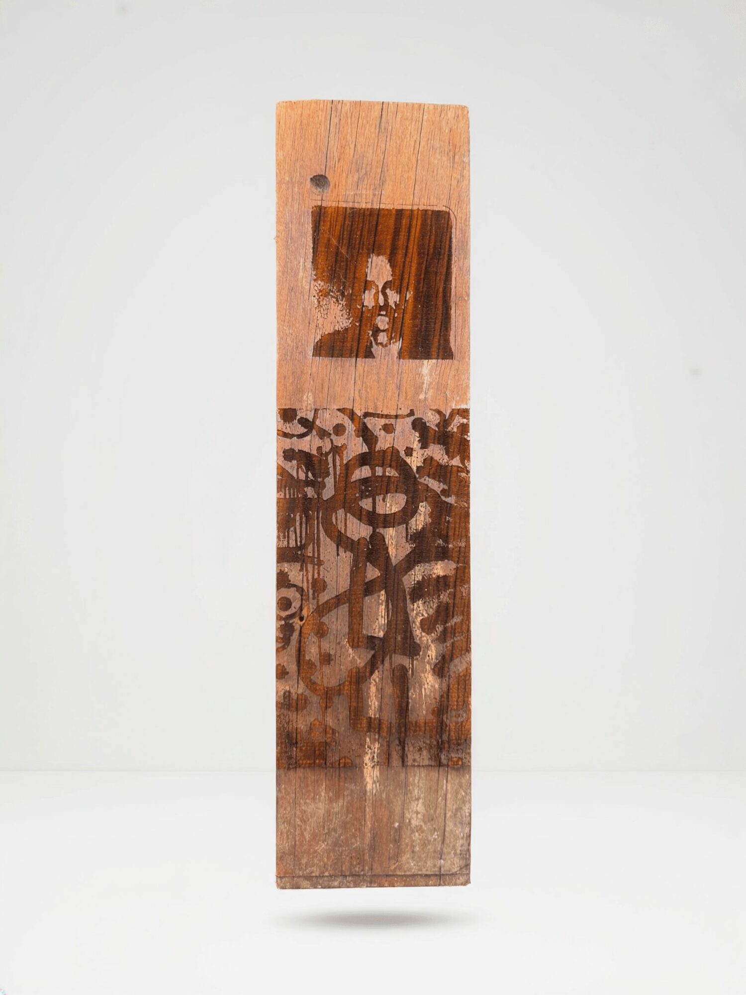 A tall wooden plank with a small photo of a person at the top and abstract symbols below, standing upright.