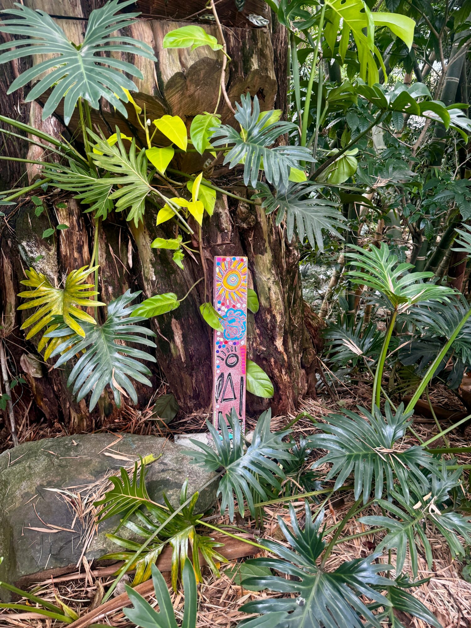 Colorful painted wooden post with geometric and floral designs stands among lush green tropical plants and trees.