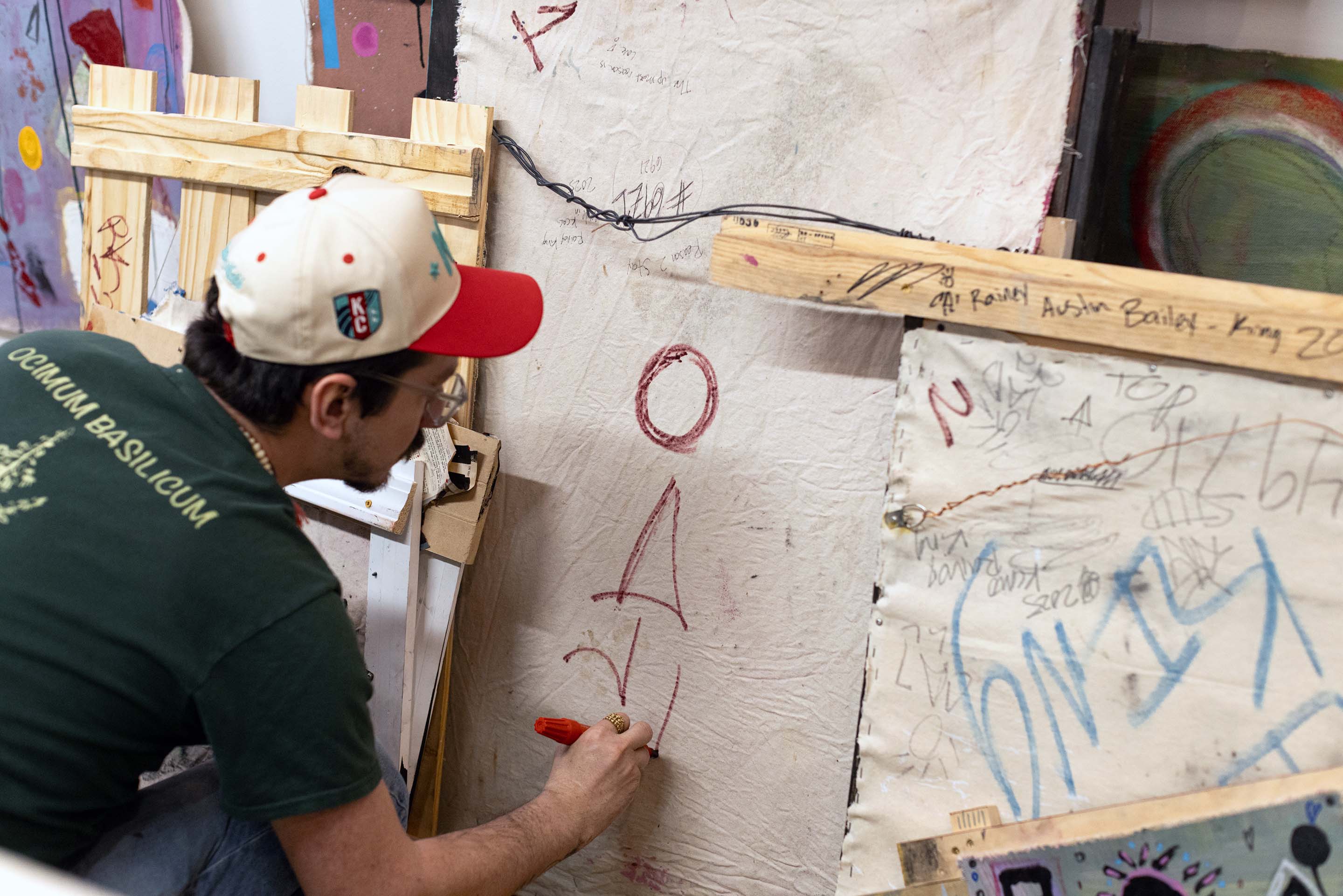 Person drawing on a whiteboard with red marker, surrounded by various objects and graffiti, in a cluttered room.