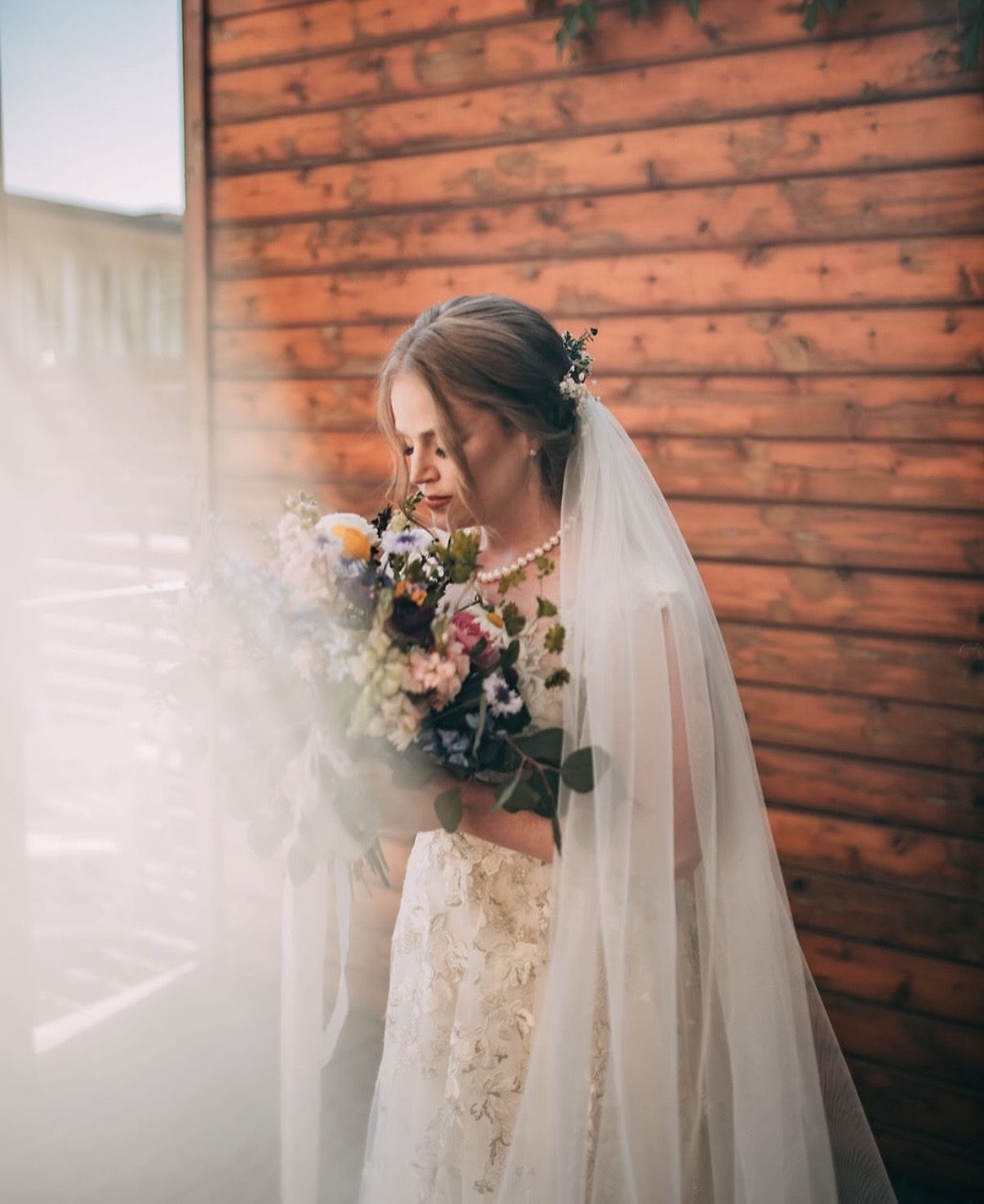 Bride with a veil holding a bouquet, standing against a wooden wall, looking down at flowers, soft lighting.