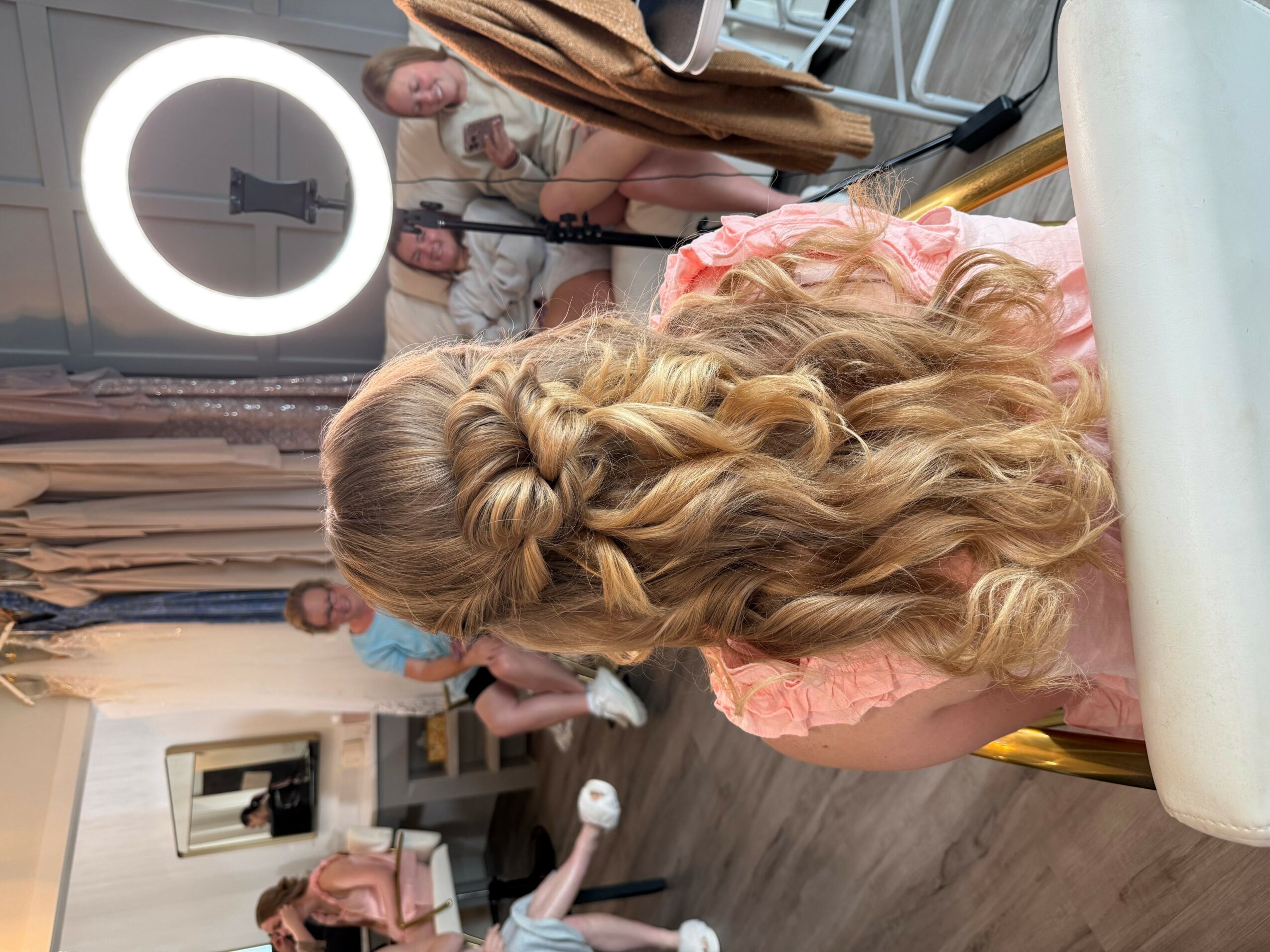 Person with long, curly blonde hair styled in an intricate braid, sitting at a table in a room with people and a ring light.