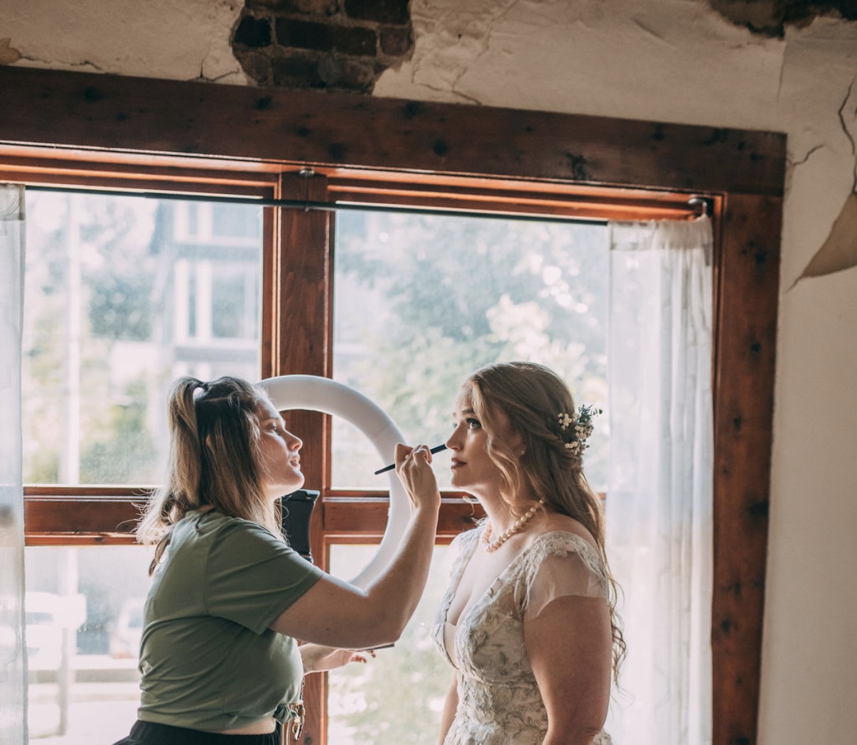 Two women, one applying makeup to the other, standing near a window with curtains, sunlight streaming in.