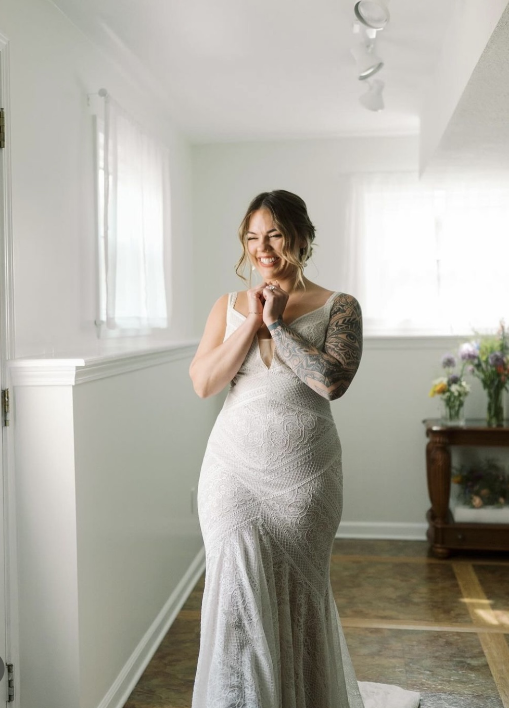 Woman in wedding dress smiling with hands clasped, standing in a bright room with windows and a wooden table with flowers.