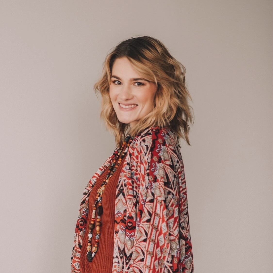 Young woman with wavy blonde hair smiling, wearing a patterned red and pink top and beaded necklace, against a plain background.