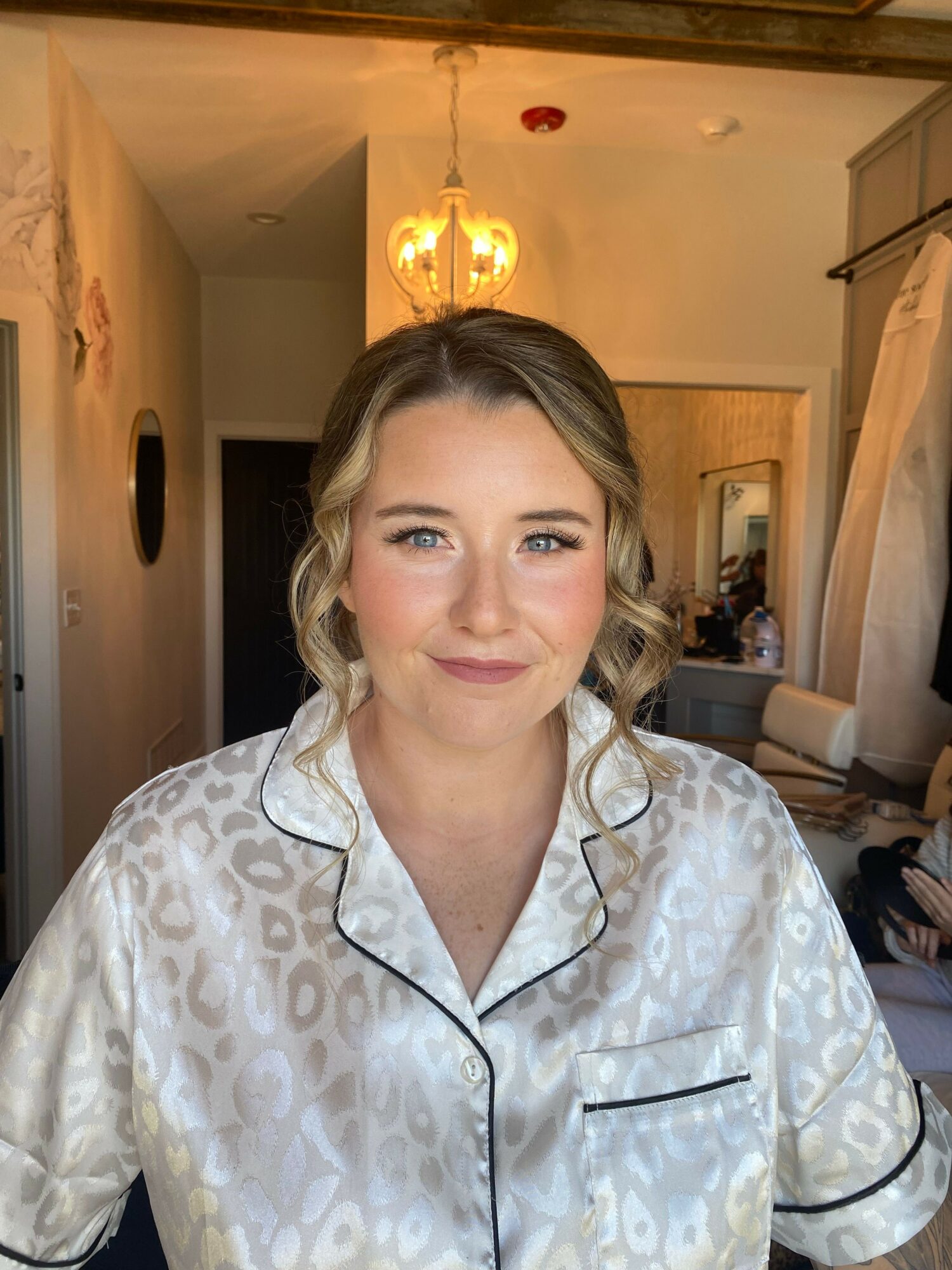 Woman with blonde hair in a satin patterned shirt, smiling, in a warmly lit room with a lamp and mirror.