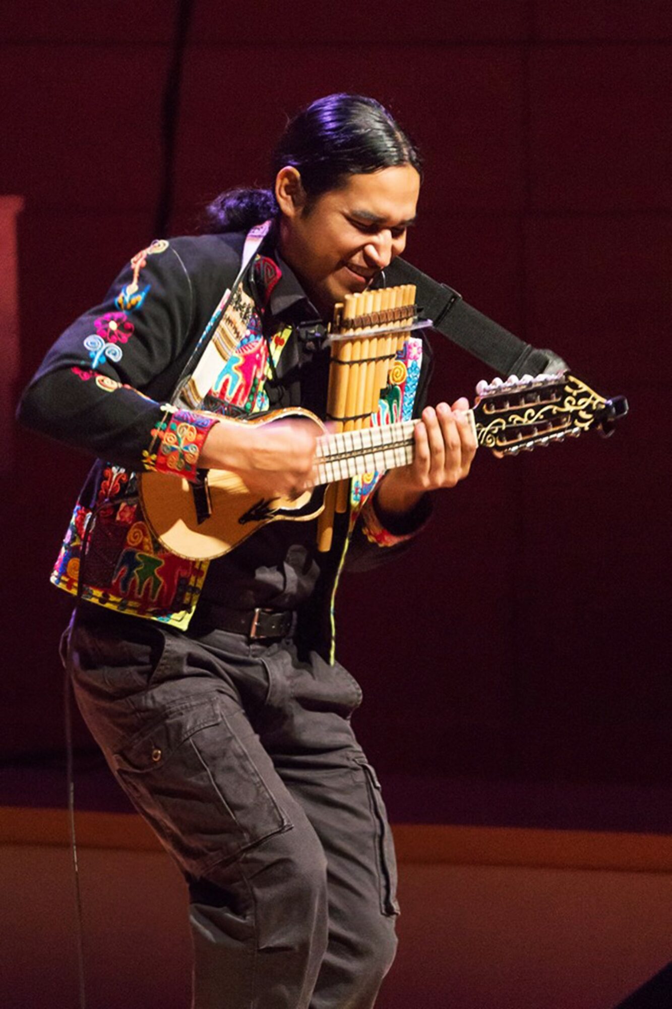 Person playing a small stringed instrument with a joyful expression, wearing a colorful jacket, on stage.