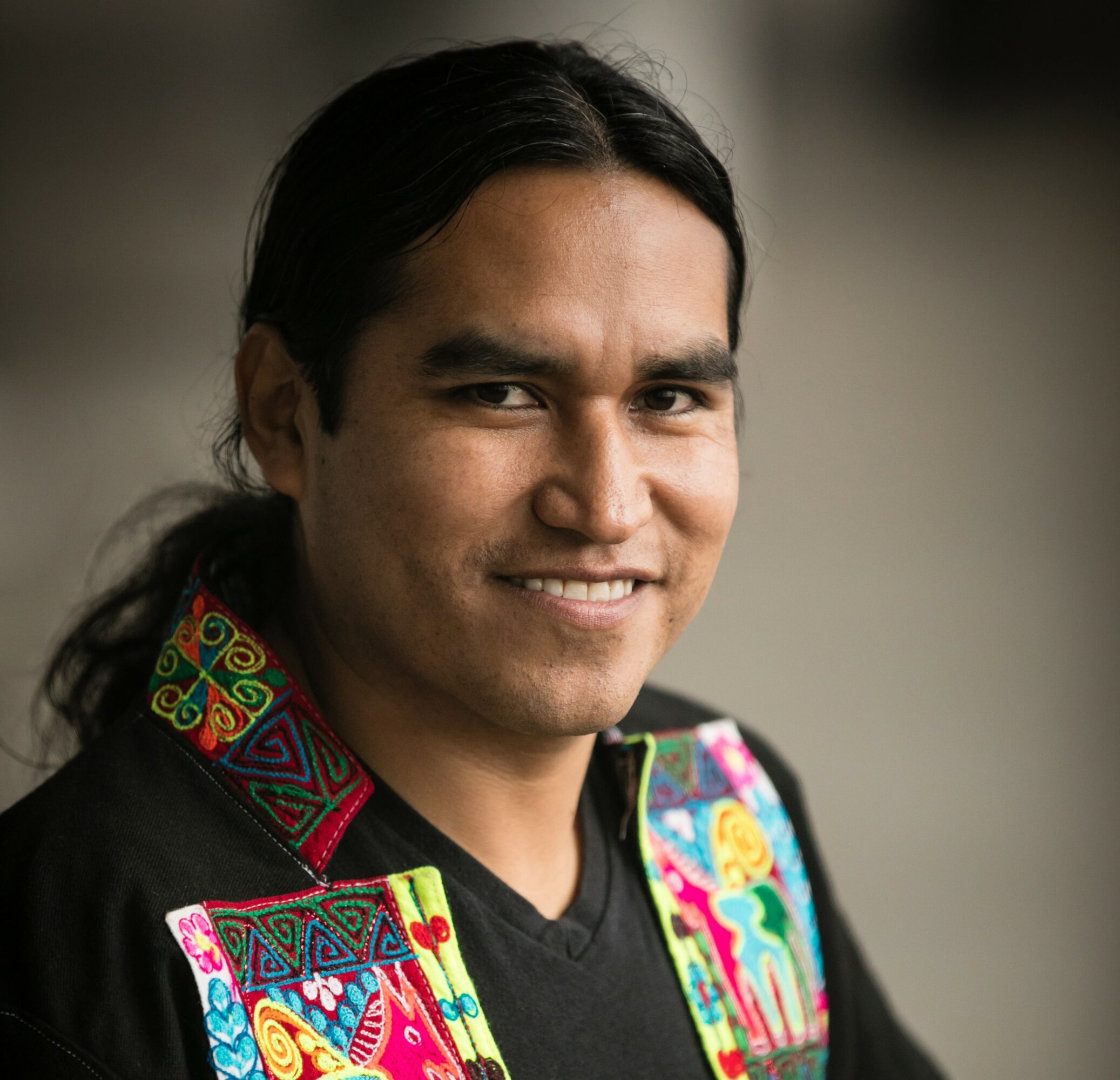 Portrait of a person with long dark hair, smiling, wearing a black shirt with colorful embroidered patterns on the shoulders and collar.
