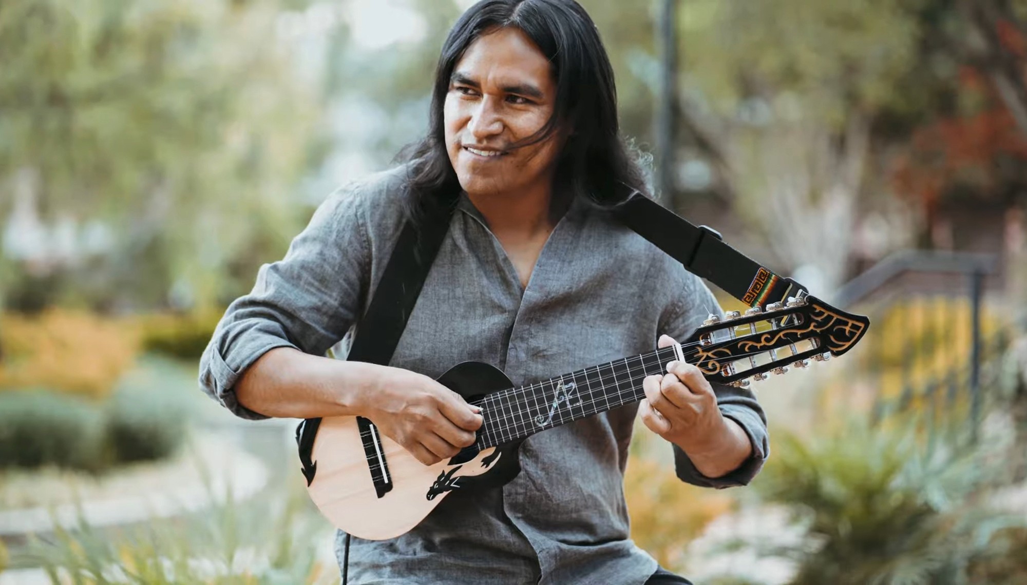 Person with long dark hair playing a small guitar outdoors, smiling, with trees and a fence in the background.