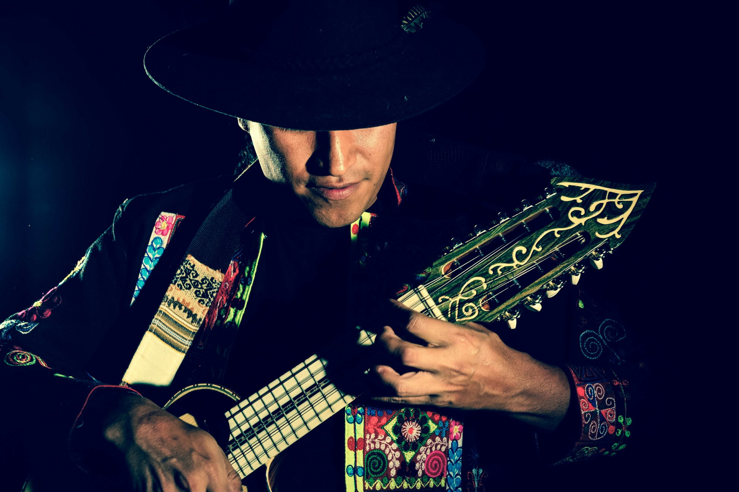 Person wearing a wide-brimmed hat playing a stringed instrument with decorative patterns, dark background.