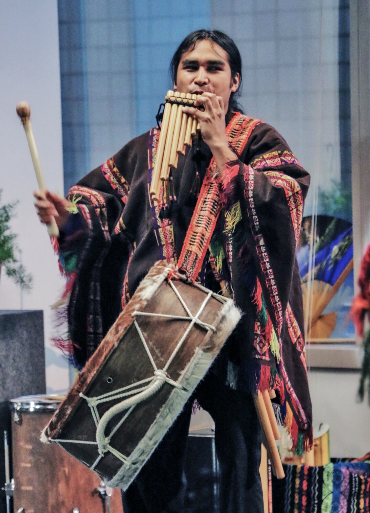 Person playing pan flute, wearing colorful traditional clothing, holding drum and drumsticks, outdoors with modern building background.