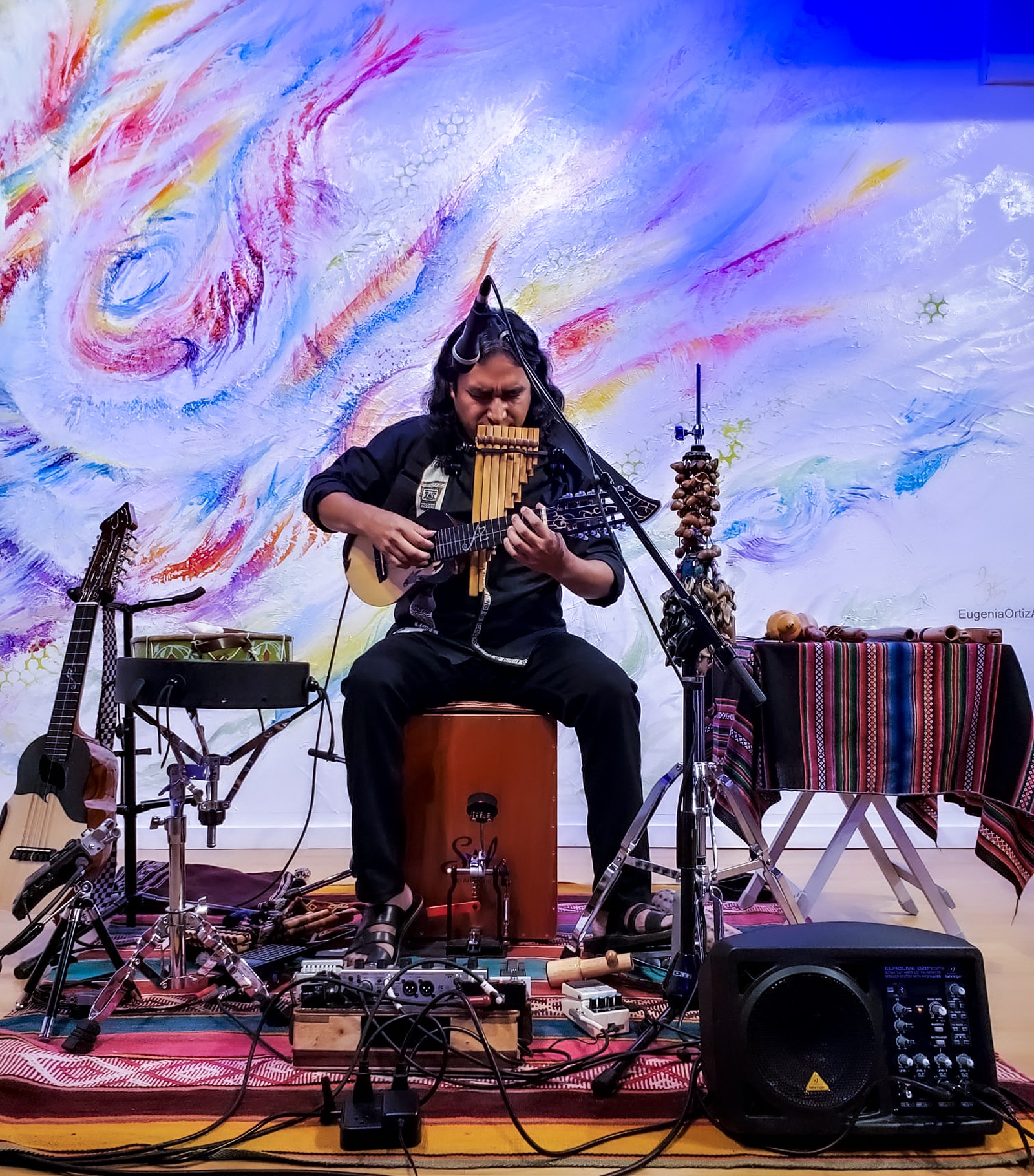 Person playing a musical instrument on stage with colorful abstract background, surrounded by musical equipment and a table with a striped cloth.