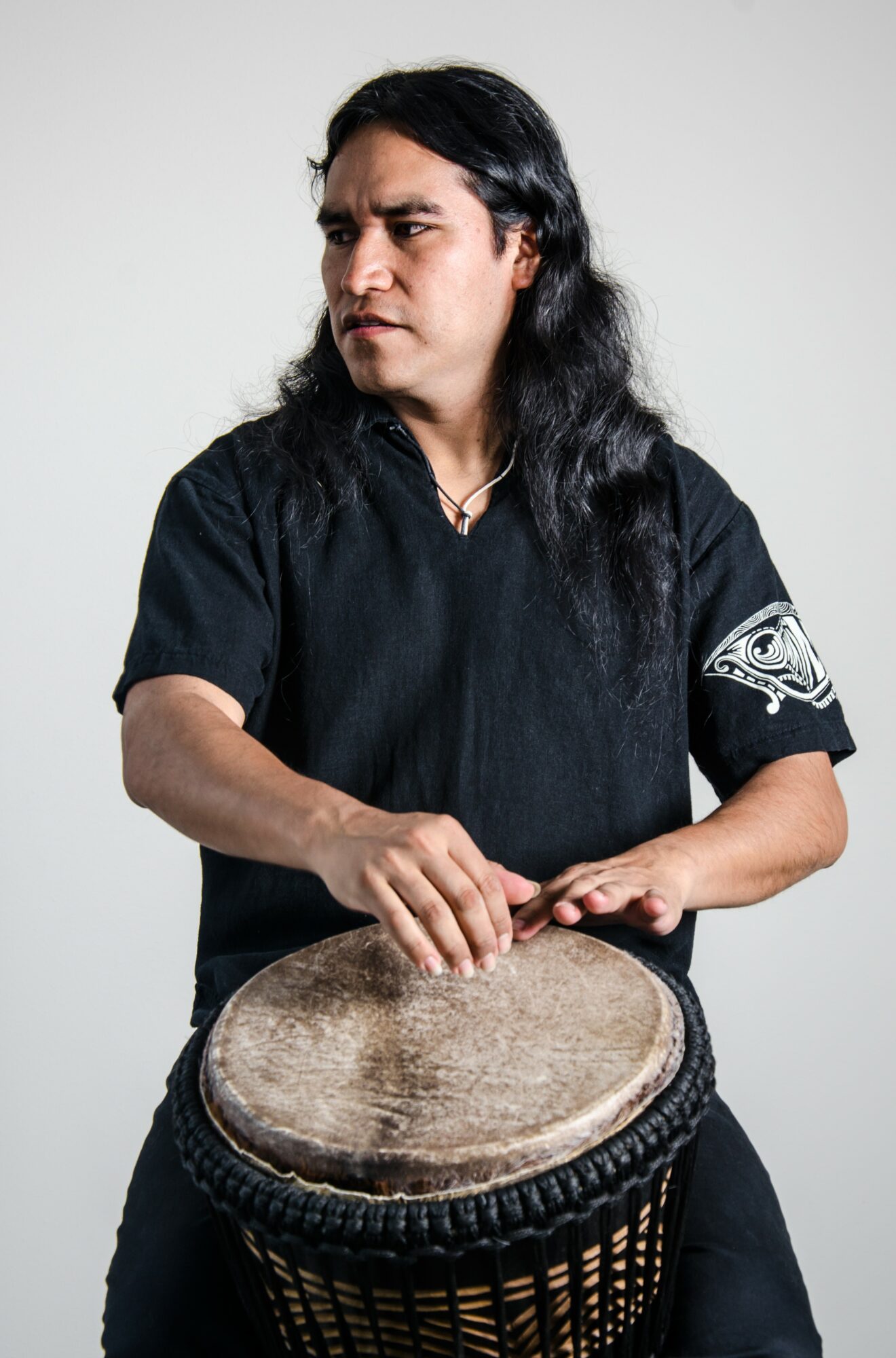 Person with long dark hair playing a drum, wearing a black shirt with a white design on the sleeve, against a plain background.