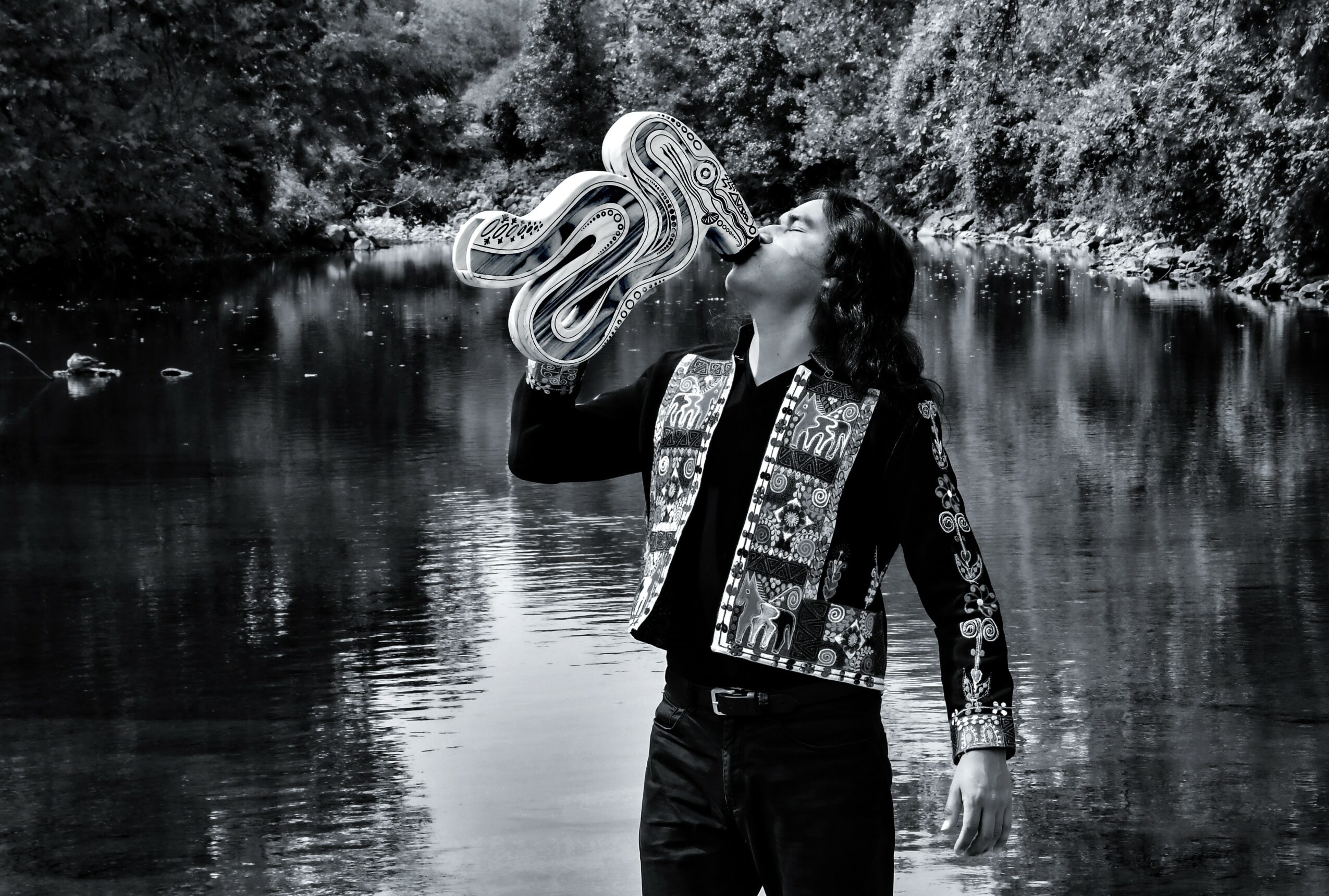 Person drinking from a large, coiled object near a river with trees in the background, in black and white.